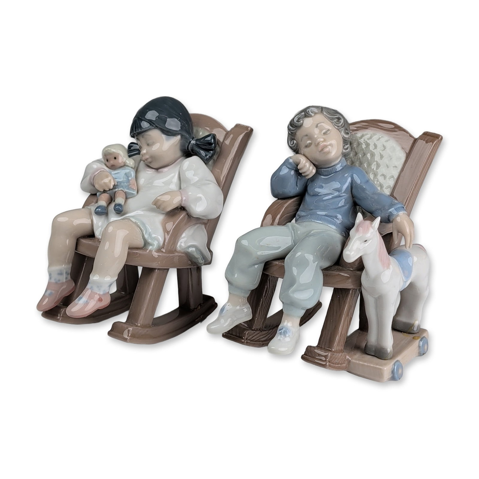 Two Lladro Porcelain Figurines, Naptime 01005448 & All Tuckered Out 01005846: Porcelain figurines featuring two sleeping children in rocking chairs. One figurine depicts a boy in a blue sweater and light blue trousers holding a white rocking horse with blue wheels. The other de