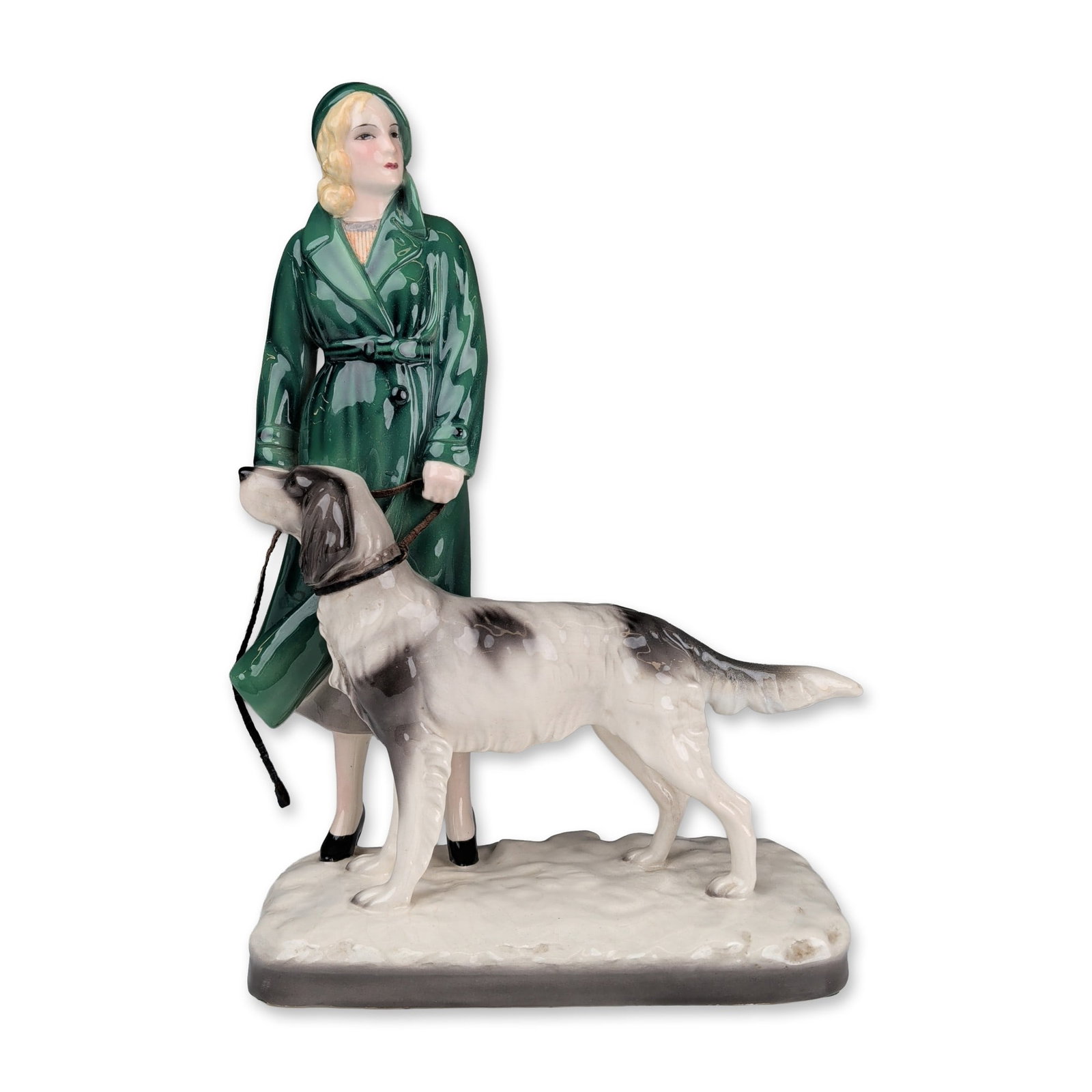 Goldscheider by Stephan Dakon Art Deco Lady with Setter Figurine, 6751: The polychrome glazed work depicts a woman in a high-gloss emerald green trench coat and matching cloche hat walking a white and black spotted setter. The figures are positioned on an off-white textur