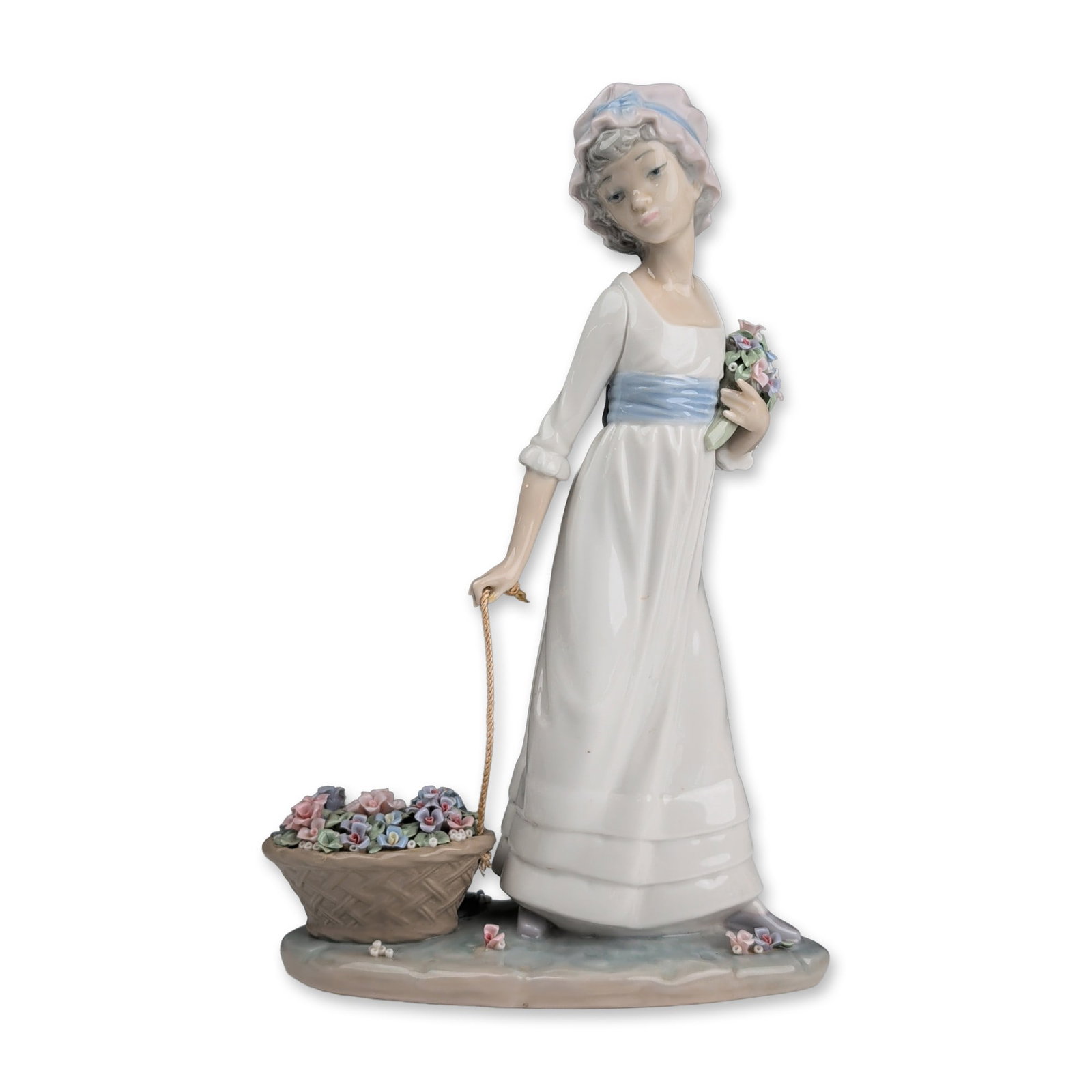 Lladro Porcelain Figurine, Wildflower 01005030: Lladro porcelain figurine sculpted by Vincente Martinez. The piece depicts a young woman in a white empire-waist gown with a blue sash and matching bonnet, pulling a woven basket of flowers on a wheel