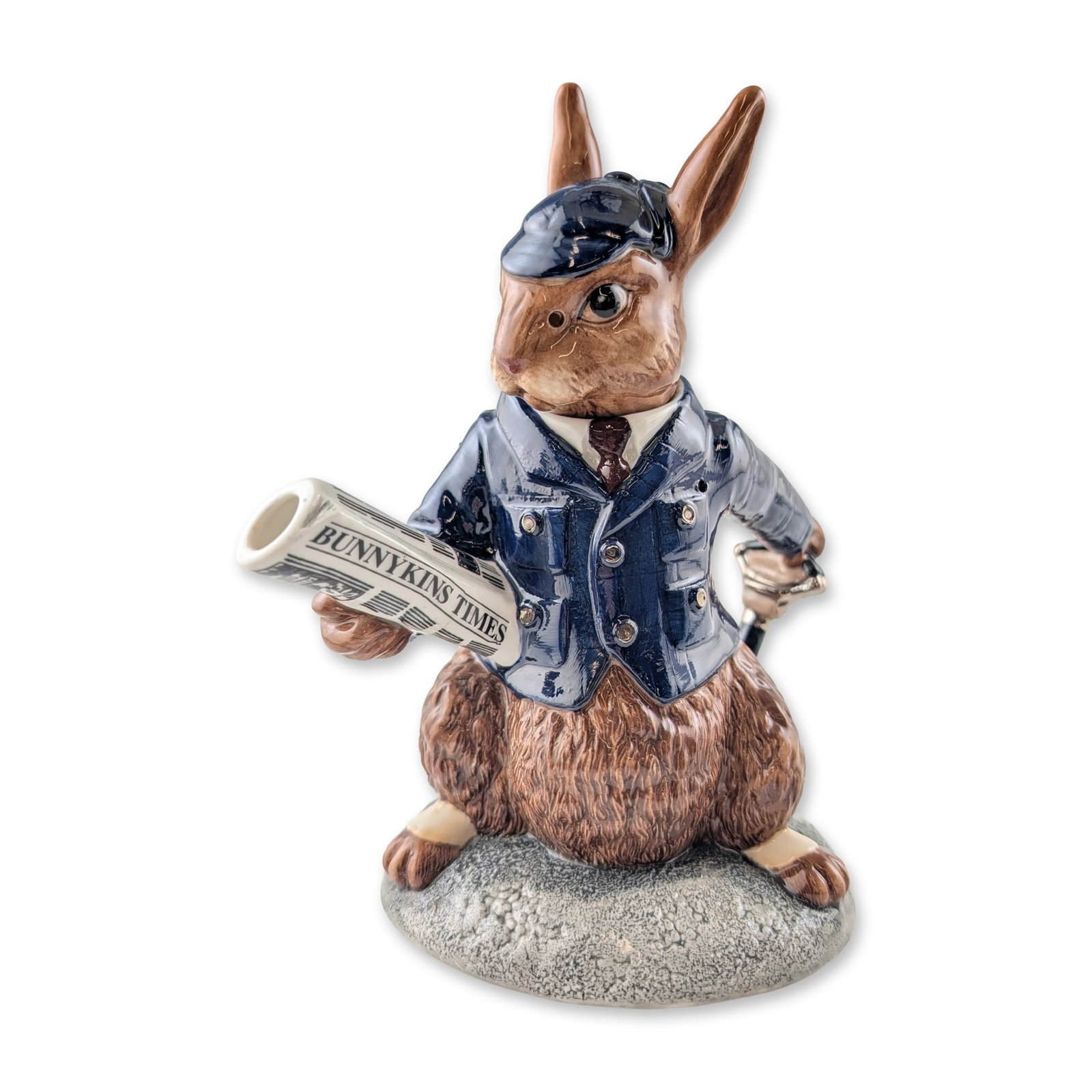 Royal Doulton Bunnykins Prototype Teapot, Lord of the Manor DBD1: The figure is modeled in a navy-blue double-breasted Norfolk-style jacket with a white collar and burgundy tie, wearing a matching blue flat cap. A rolled newspaper titled Bunnykins Times serves as th