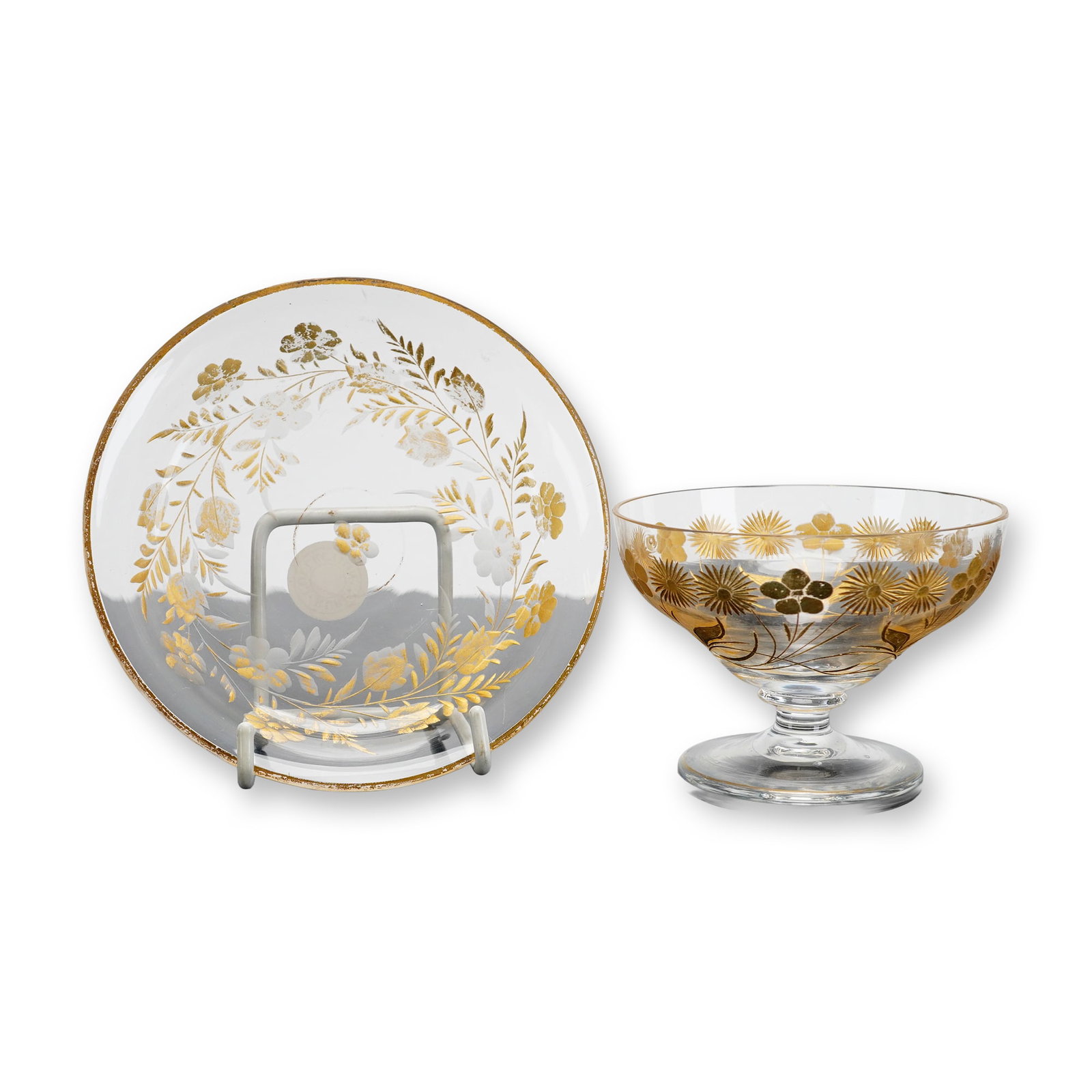 Two Clear and Gilded Glass Tableware Items: This pair of clear glass items consists of a footed dessert bowl and a matching saucer. Both pieces are decorated with ornate gilded floral and foliate motifs, including stylized sunburst flowers and 