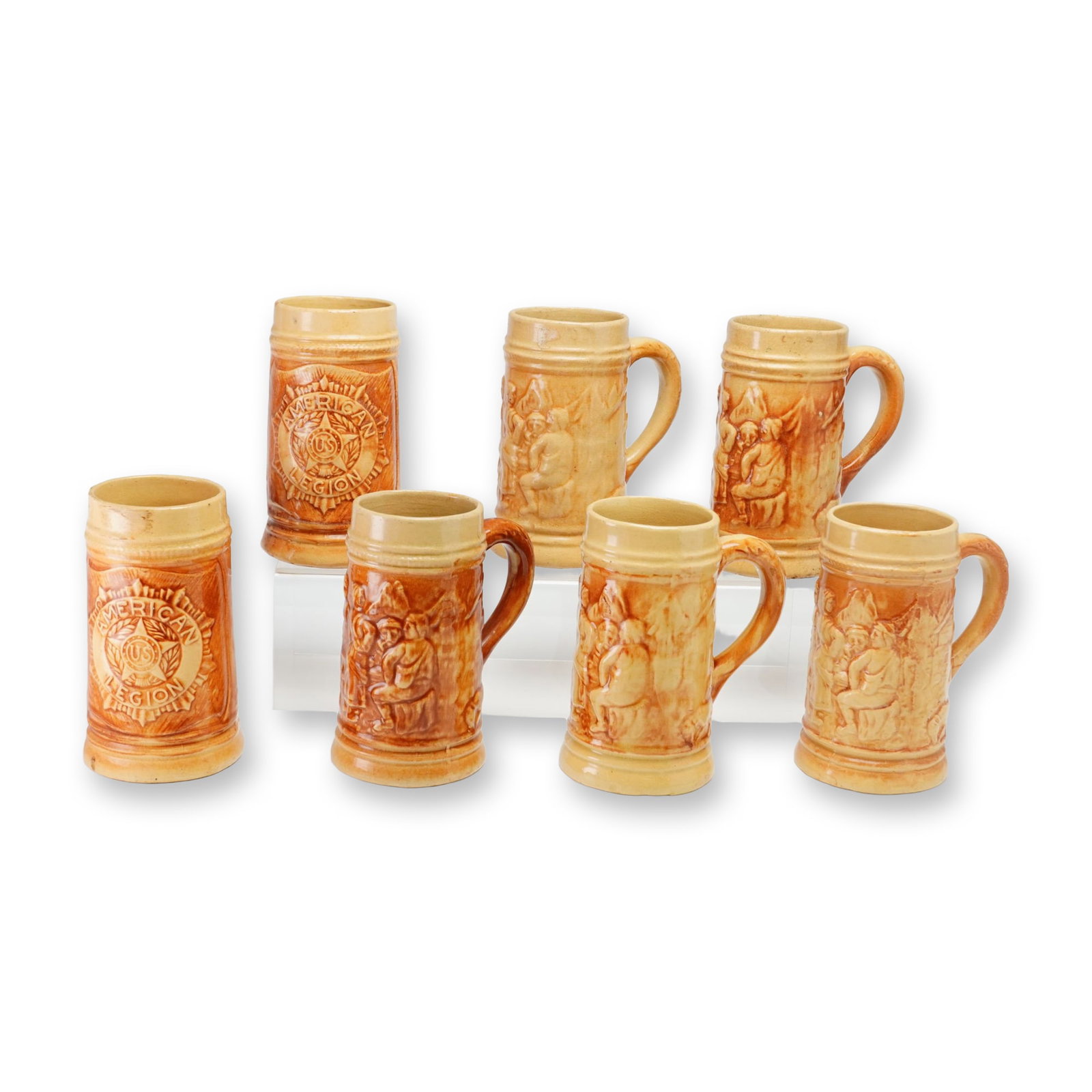 Set of Seven Hull Pottery Yellowware 1920s Steins, American Legion and Tavern Patterns: A set of seven stoneware mugs manufactured by Hull Pottery in Zanesville, Ohio. The collection features two distinct relief-molded designs. Two mugs display the American Legion emblem, which includes 