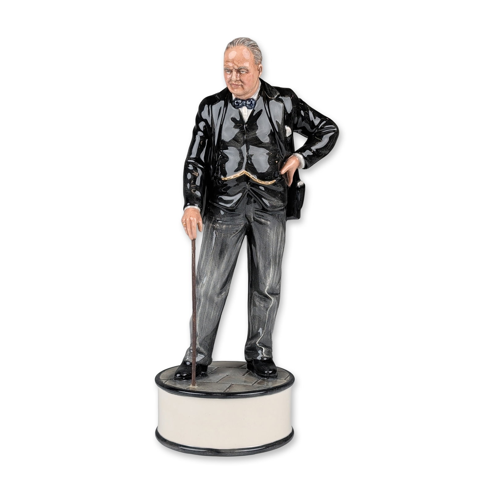 Royal Doulton Prototype Porcelain Figurine, Winston Churchill: Glazed porcelain figurine depicting Sir Winston Churchill in formal attire, including a black jacket, gray pinstripe trousers, and blue bow tie. The figure stands on a circular cobblestone patterned b