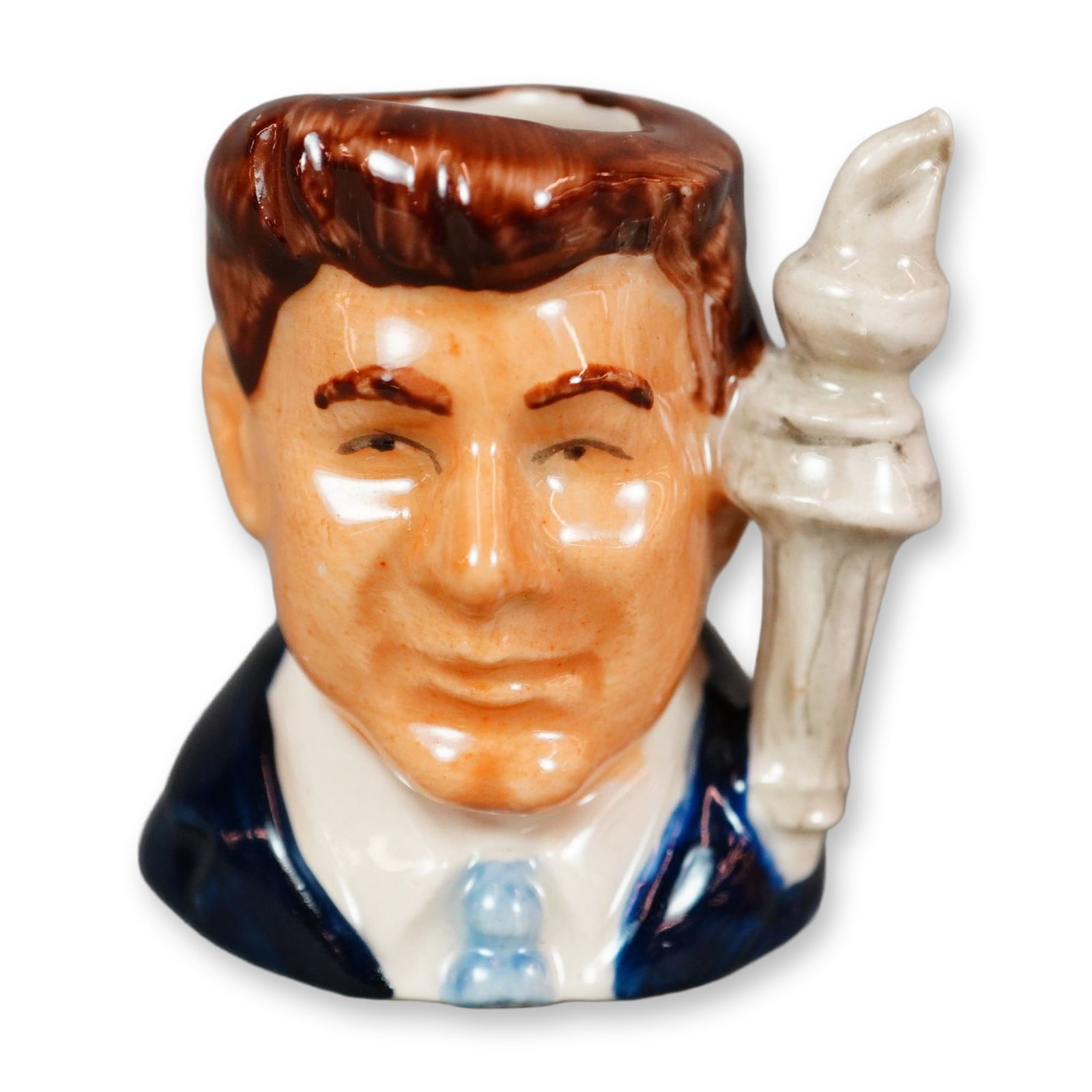 Royal Doulton Prototype Colorway Tiny Character Jug, John F. Kennedy: Figural jug depicting John F. Kennedy, shown wearing a dark blue coat with a light blue tie and brown hair. Handle modeled as a gray flaming torch. Features Royal Doulton backstamp. Dimensions: 1.5"L 