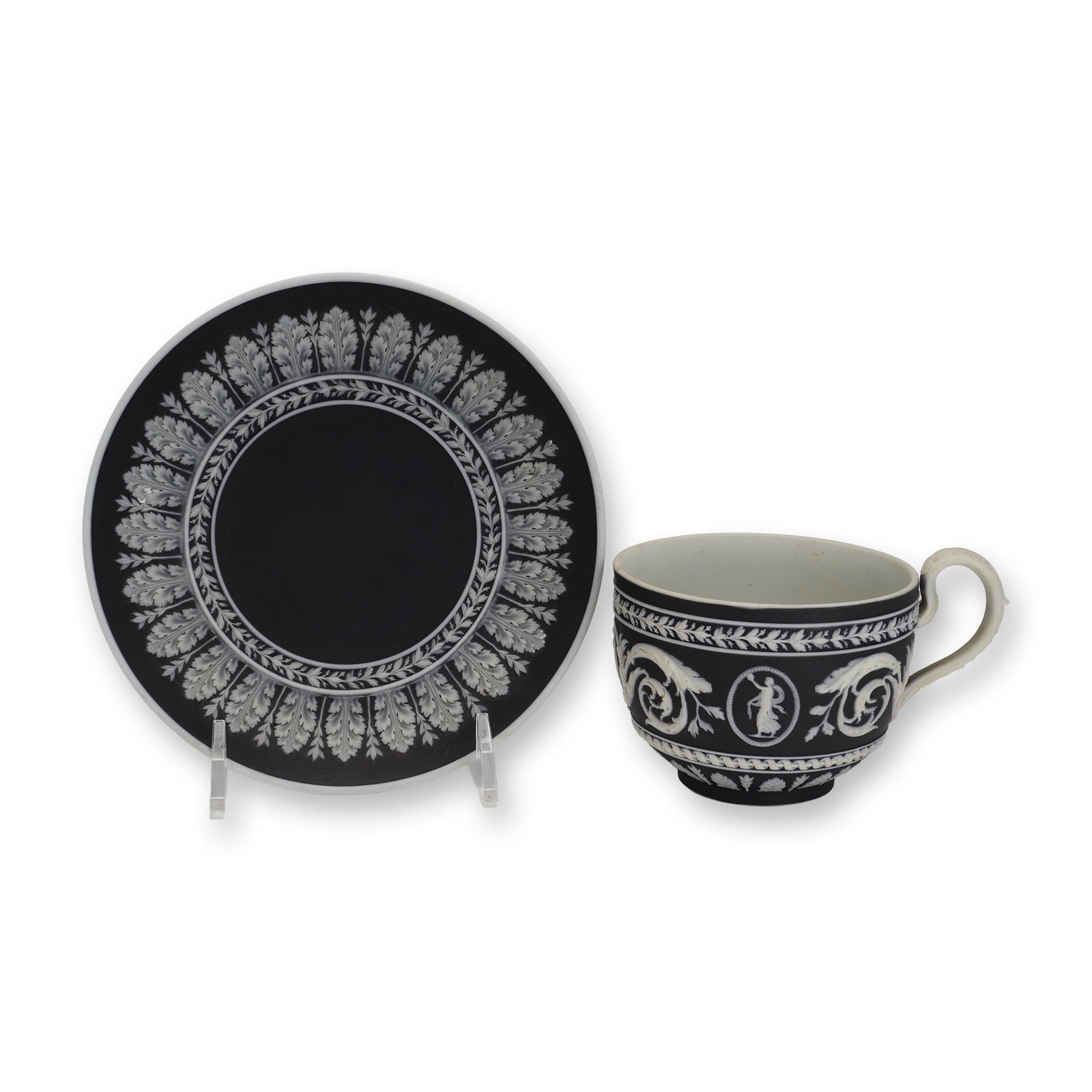 2pc Wedgwood Black and White Jasperware Cup and Saucer: A two-piece Wedgwood jasperware set comprising a tea cup and a saucer. The set is crafted in black dip jasperware, featuring a matte black surface with applied white relief sprigged neoclassical ornam