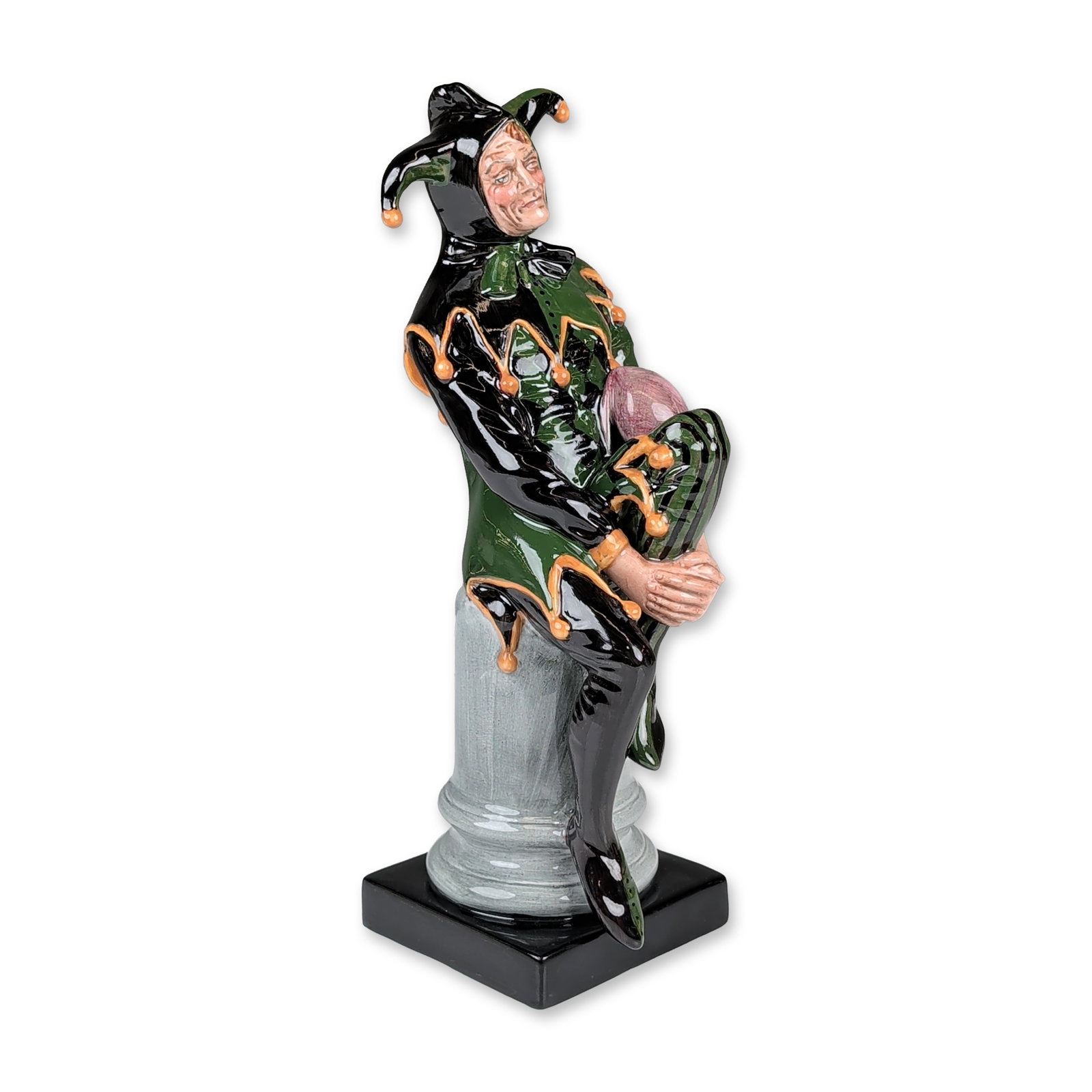 Royal Doulton Colorway Figurine, The Jester: The figure is depicted seated atop a mottled gray column, wearing a green and black motley suit with orange-tipped bells and a tripartite hat. The figure stands on a black square base. Royal Doulton b