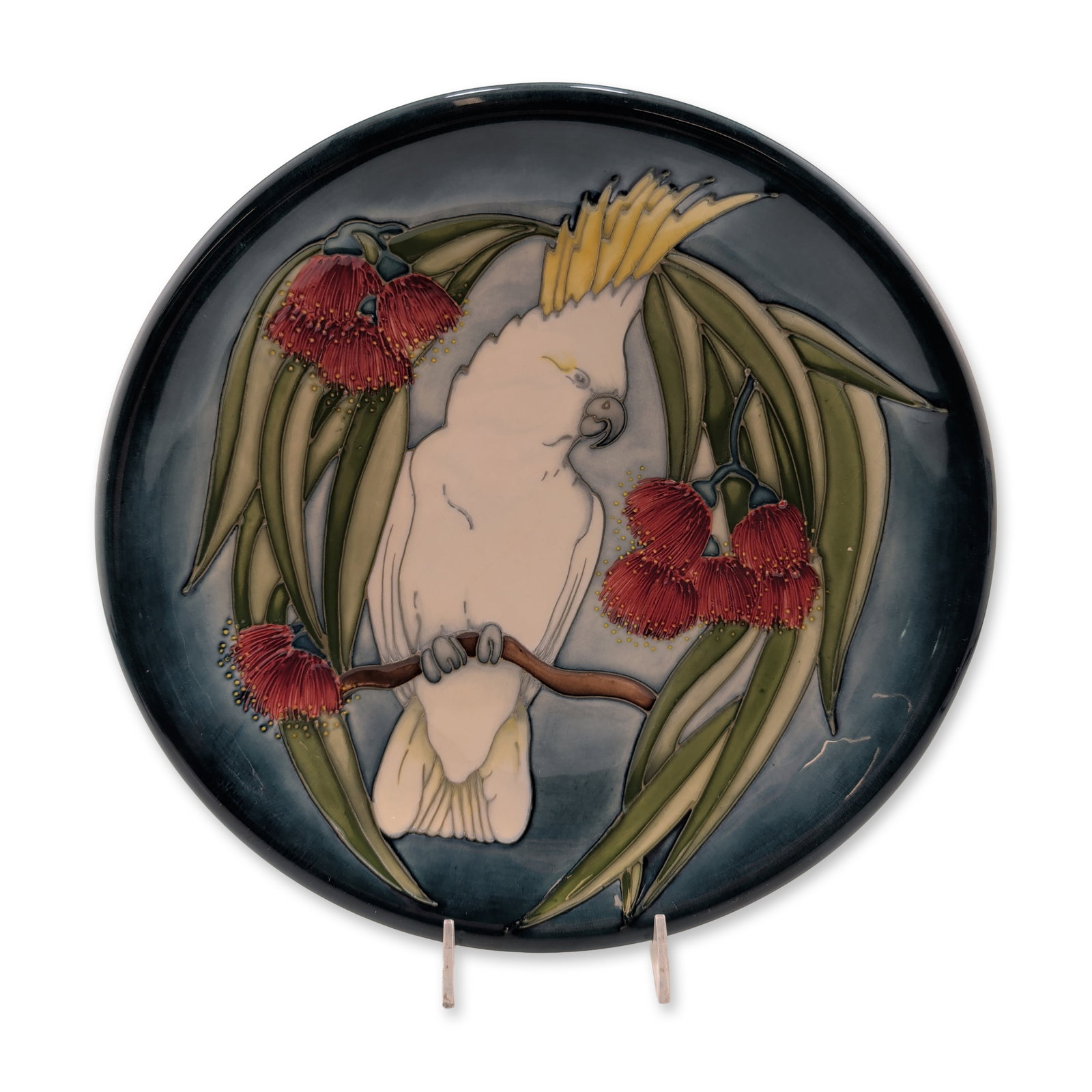 Moorcroft Pottery Trial Cockatoo Motif Cabinet Charger: Featuring a tube-lined and hand-painted design of a cockatoo among red gum blossoms and eucalyptus leaves set against a dark blue ground. The reverse features an impressed Moorcroft mark, and Trial 7-