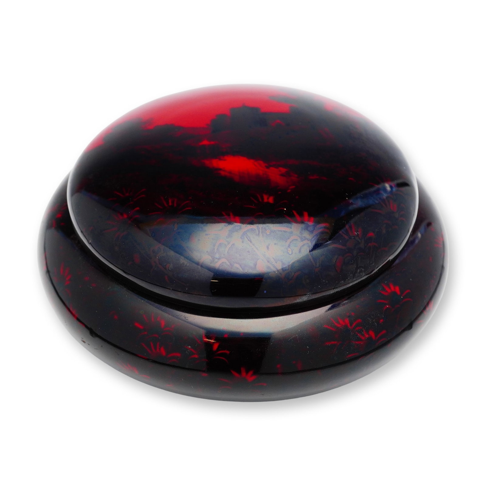 Royal Doulton Flambe Landscape Motif Round Lidded Box: A squat, round lidded box finished in a red-fired glaze, decorated with a dark-toned landscape scene encircling the body, accented by finely detailed floral motifs. The lid continues the floral decora