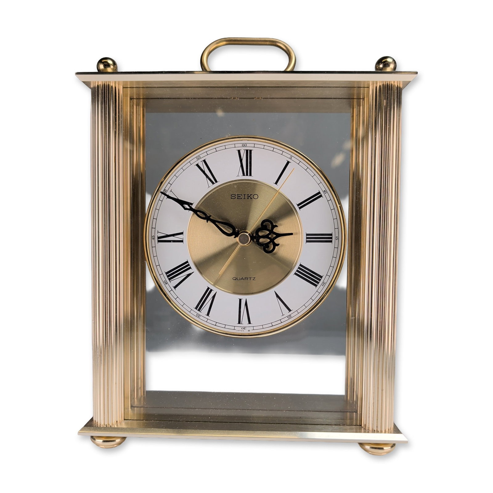 Seiko Gold Tone Quartz Carriage Clock: Seiko carriage style mantel clock in a gold tone metal and glass case with fluted corner pillars and a swinging top handle. The circular dial is configured with a white chapter ring, black Roman numer