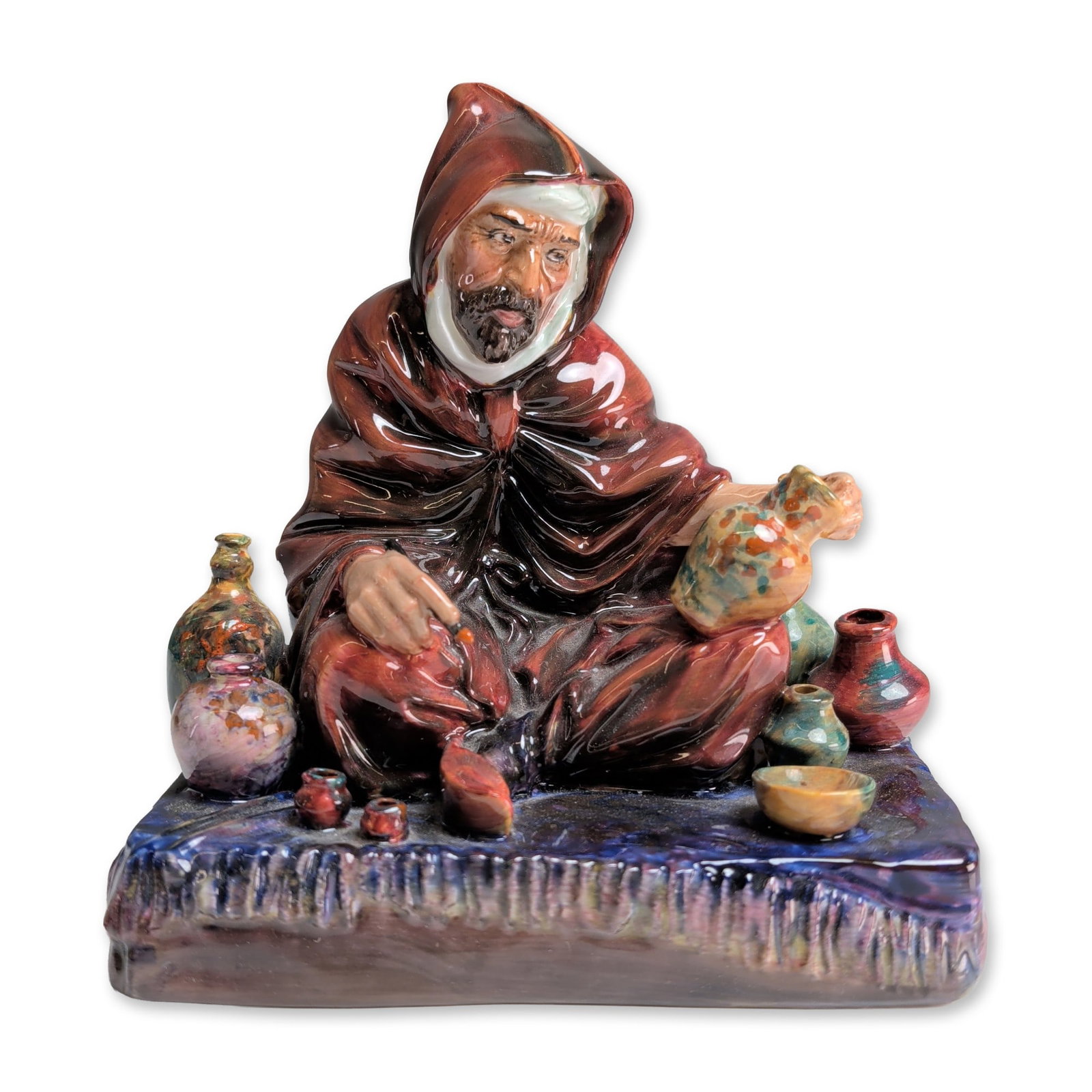 Royal Doulton Bone China Figurine, The Potter HN1493: Designed by Charles Noke. The figure is depicted seated in a hooded deep red robe and white turban, surrounded by various miniature ceramic vessels on a textured purple and blue base. The piece featur