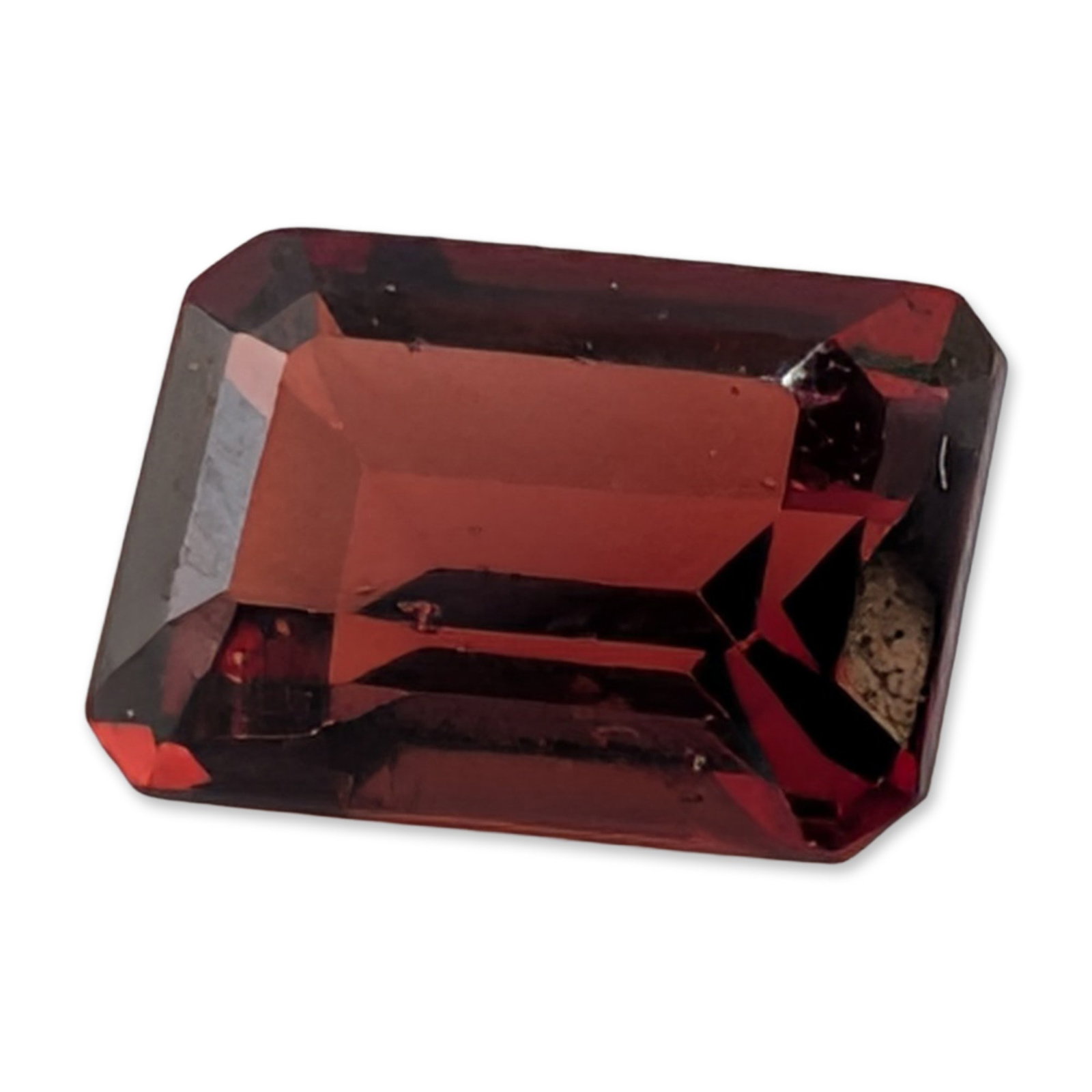 Emerald Cut Red Garnet Loose Gemstone: Loose emerald cut red gemstone appearing to be a variety of garnet. The stone displays a deep, saturated red hue with secondary brownish-orange undertones and bright red internal flashes. The rectangu