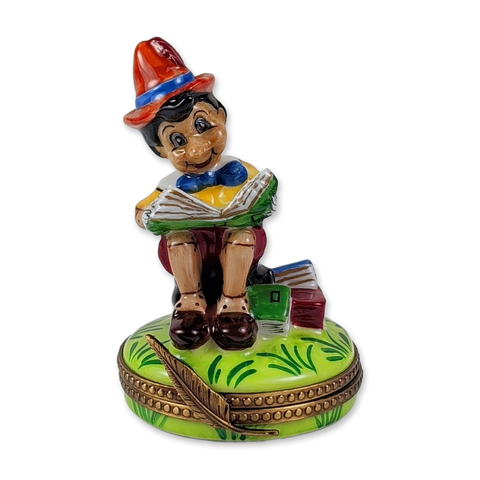 Limoges Pinocchio Porcelain Trinket Box: Limoges, France figural porcelain hinged trinket box depicting Pinocchio seated and reading a book. The figure rests on a circular green base with grass details, two books, and a metal feather quill t