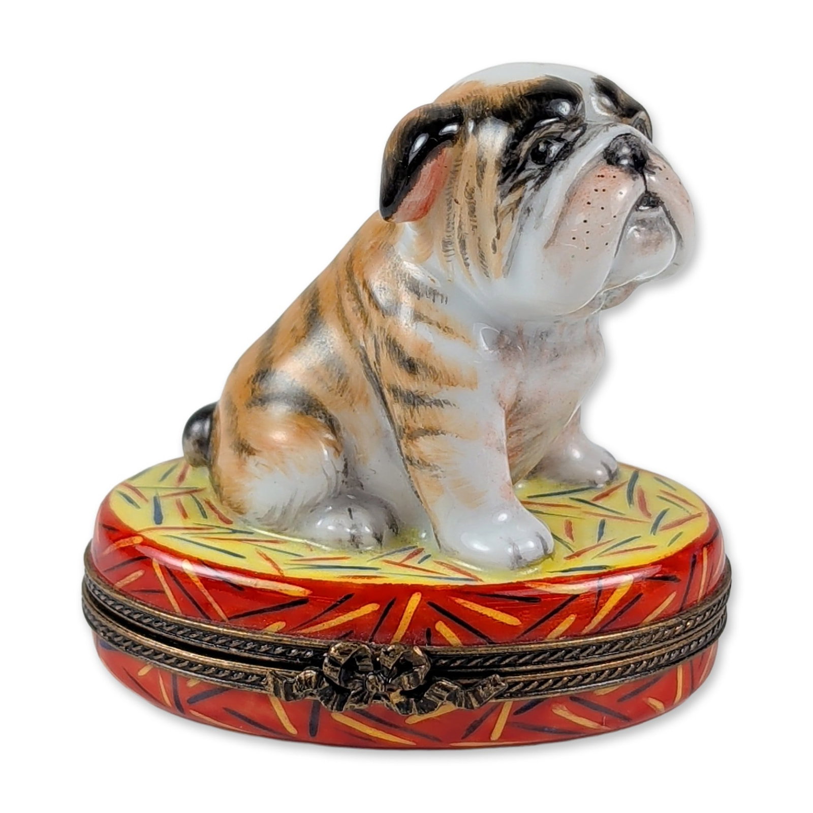 Limoges Porcelain English Bulldog Trinket Box: Limoges, France. Hand-painted porcelain figural trinket box modeled as a seated English Bulldog puppy with brindle markings on a red and yellow decorative cushion. The box includes a bronzed metal mou