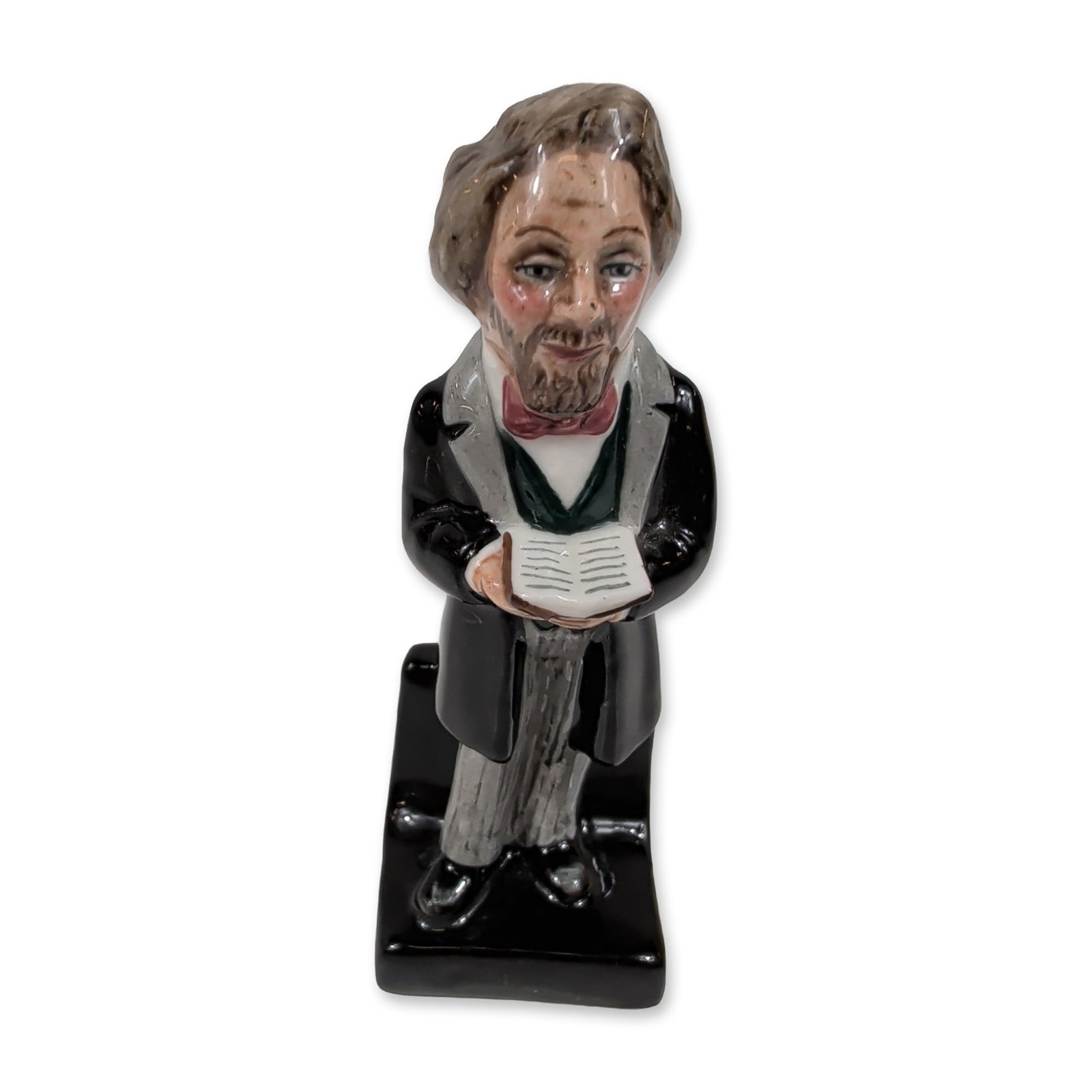 Royal Doulton Dickens Ware Porcelain Figurine, Charles Dickens HN3448: The figure is depicted standing on a black rectangular base in mid-19th-century attire, including a black frock coat, gray trousers, green waistcoat, and red bowtie, while holding an open book. Modele