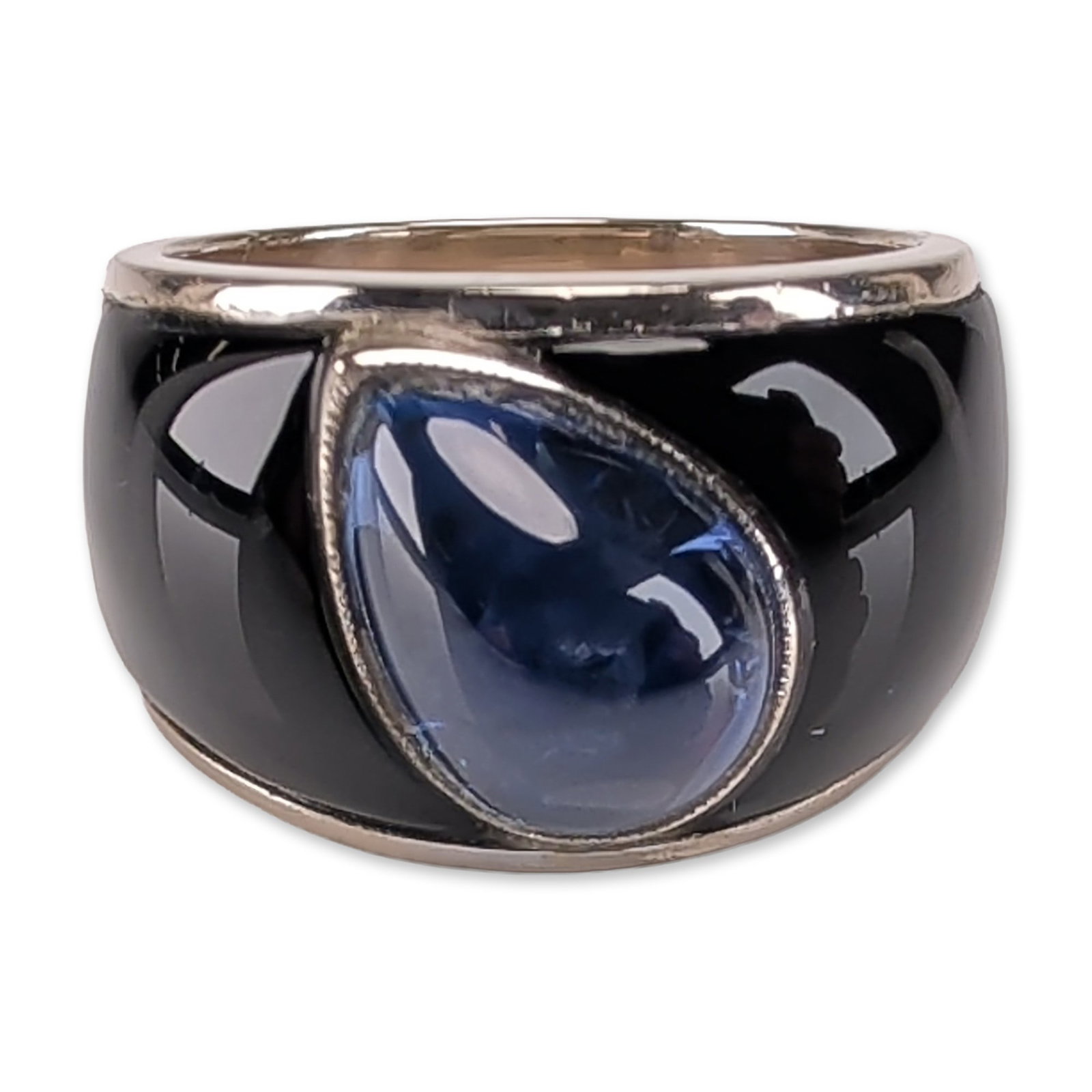 18K White Gold 4.50ct Blue Sapphire and Onyx Ring: Modernist wide band ring in 18k white gold, featuring a central pear-cut blue sapphire bezel-set at a diagonal angle, weighing 4.50 carats. The sapphire is flanked by two inlaid sections of polished b