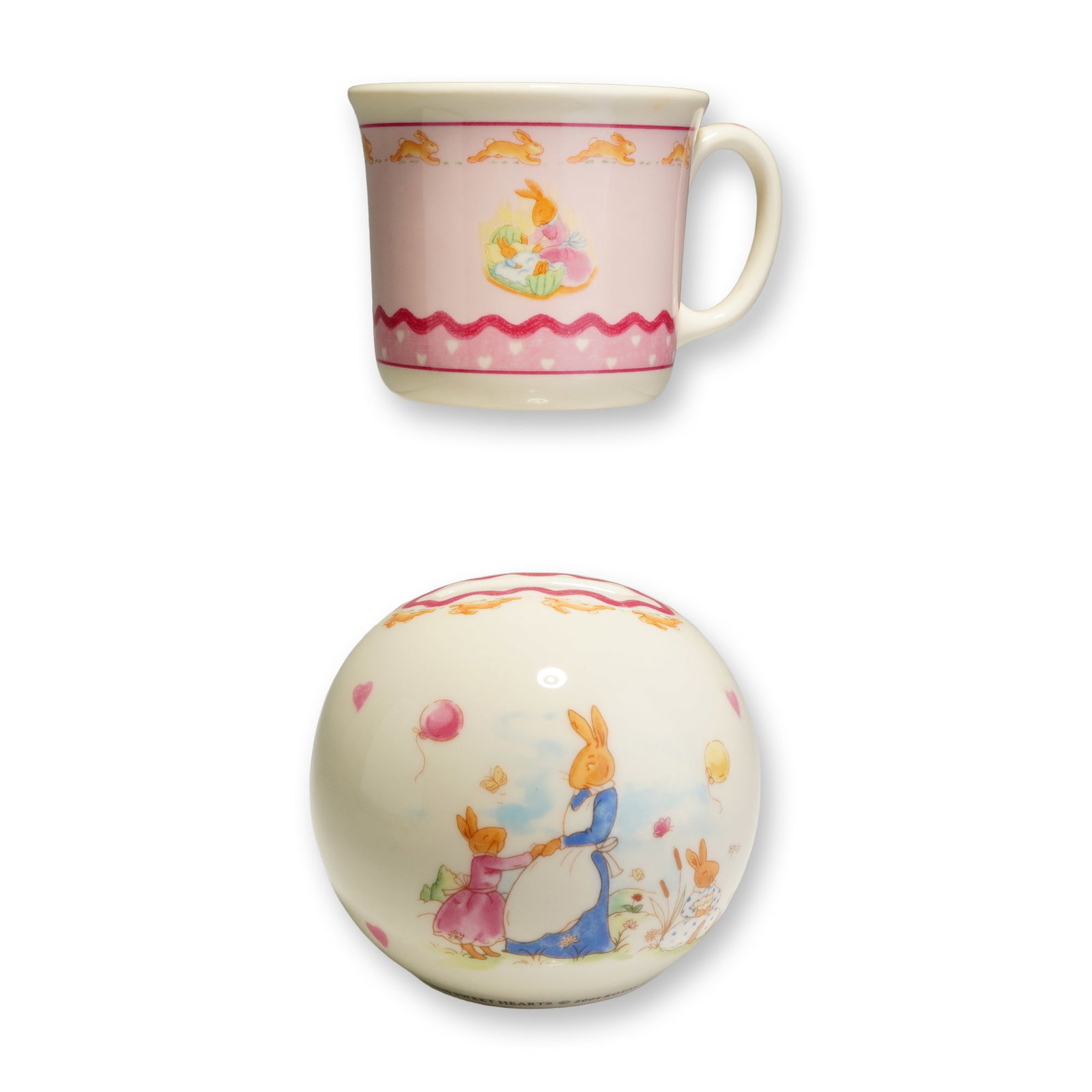 2pc Royal Doulton Bunnykins Sweet Hearts Mug and Money Bank: The handled mug and spherical money box are crafted from bone china with a cream-colored ground, while the mug features a prominent pink area as its main background. Both pieces showcase colorful illu