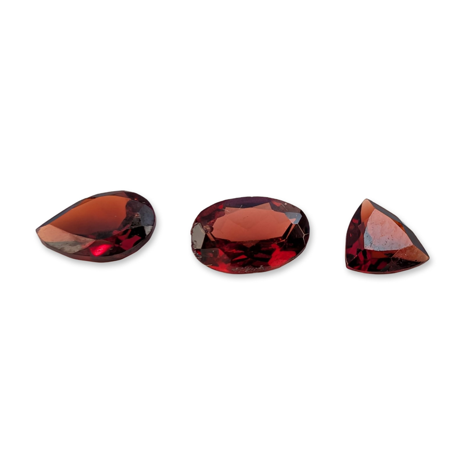 Trio of Mixed Cut Red Garnet Gemstones: Trio of loose faceted gemstones in a deep brownish-red color, which appear to be garnet. The lot includes one pear cut, one oval cut, and one trillion cut stone. Three pieces total. Dimensions: See De