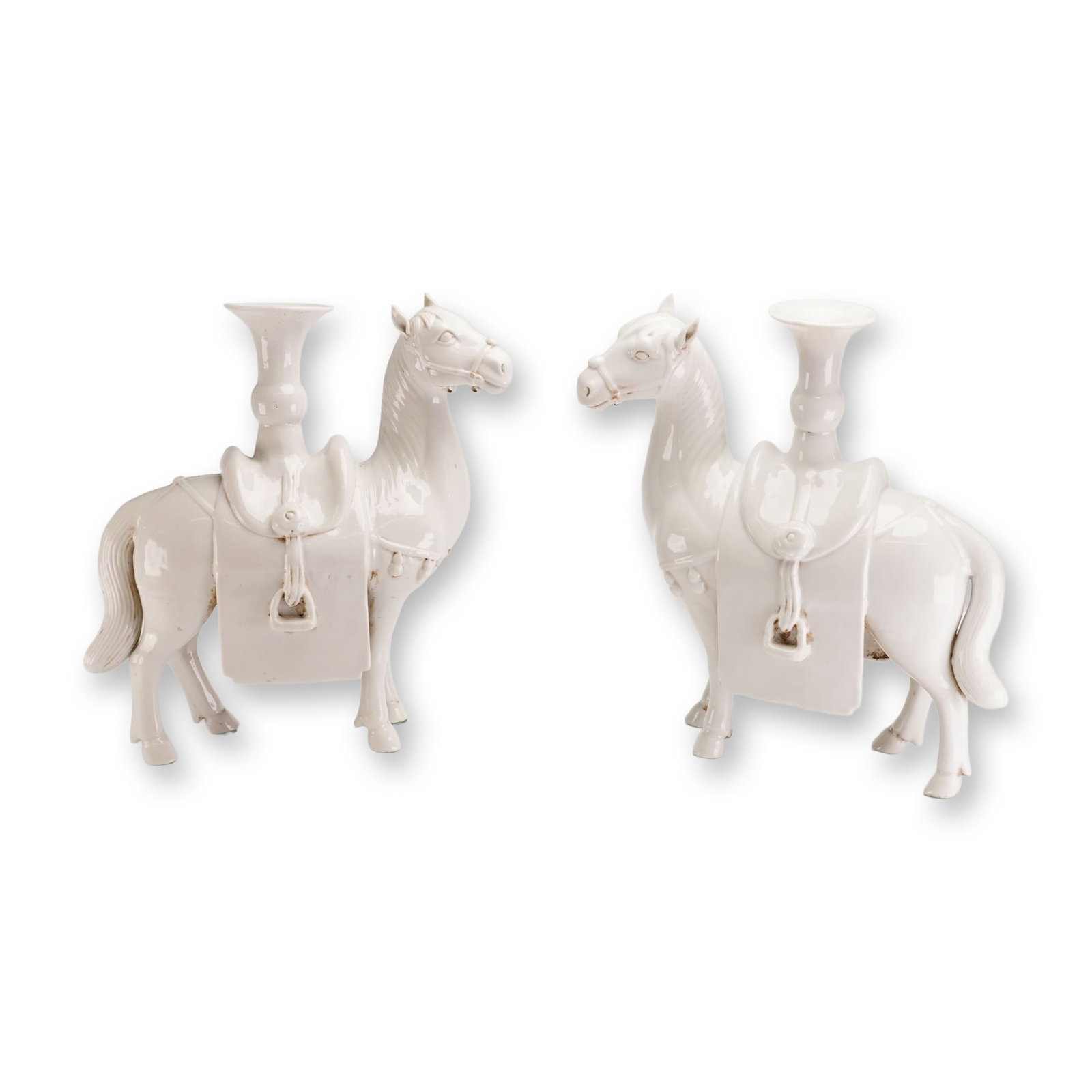 Pair of White Glazed Ceramic Figural Horse Candle Holders: Each horse is modeled in a standing position with the head turned to the side, featuring detailed molded trappings including bridles, tasseled breastplates, and saddles. A trumpet-shaped vessel with a