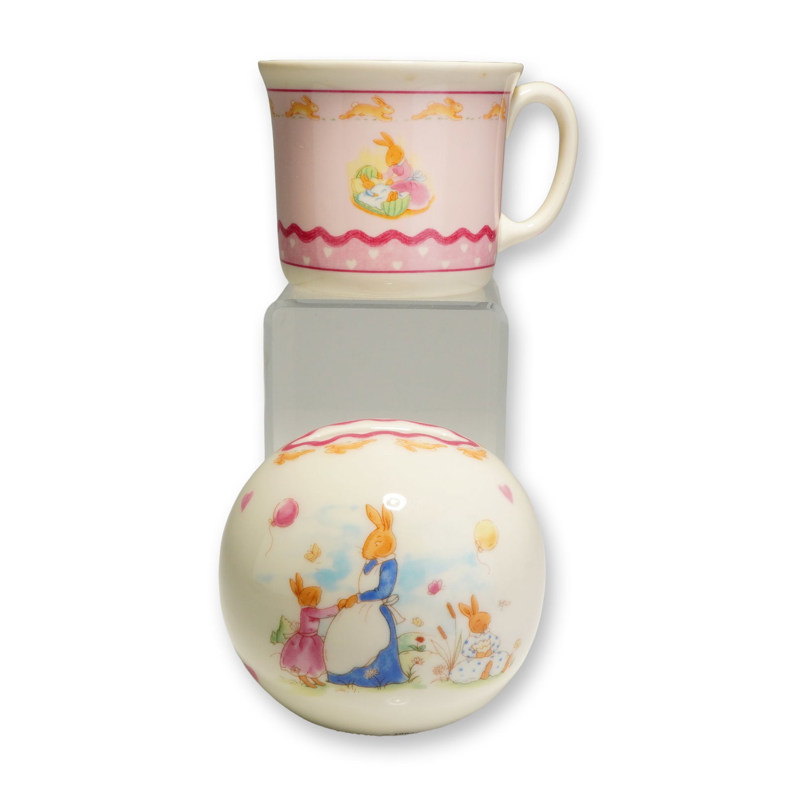2pc Royal Doulton Bunnykins Sweet Hearts Mug and Money Bank: The handled mug and spherical money box are crafted from bone china with a cream-colored ground, while the mug features a prominent pink area as its main background. Both pieces showcase colorful