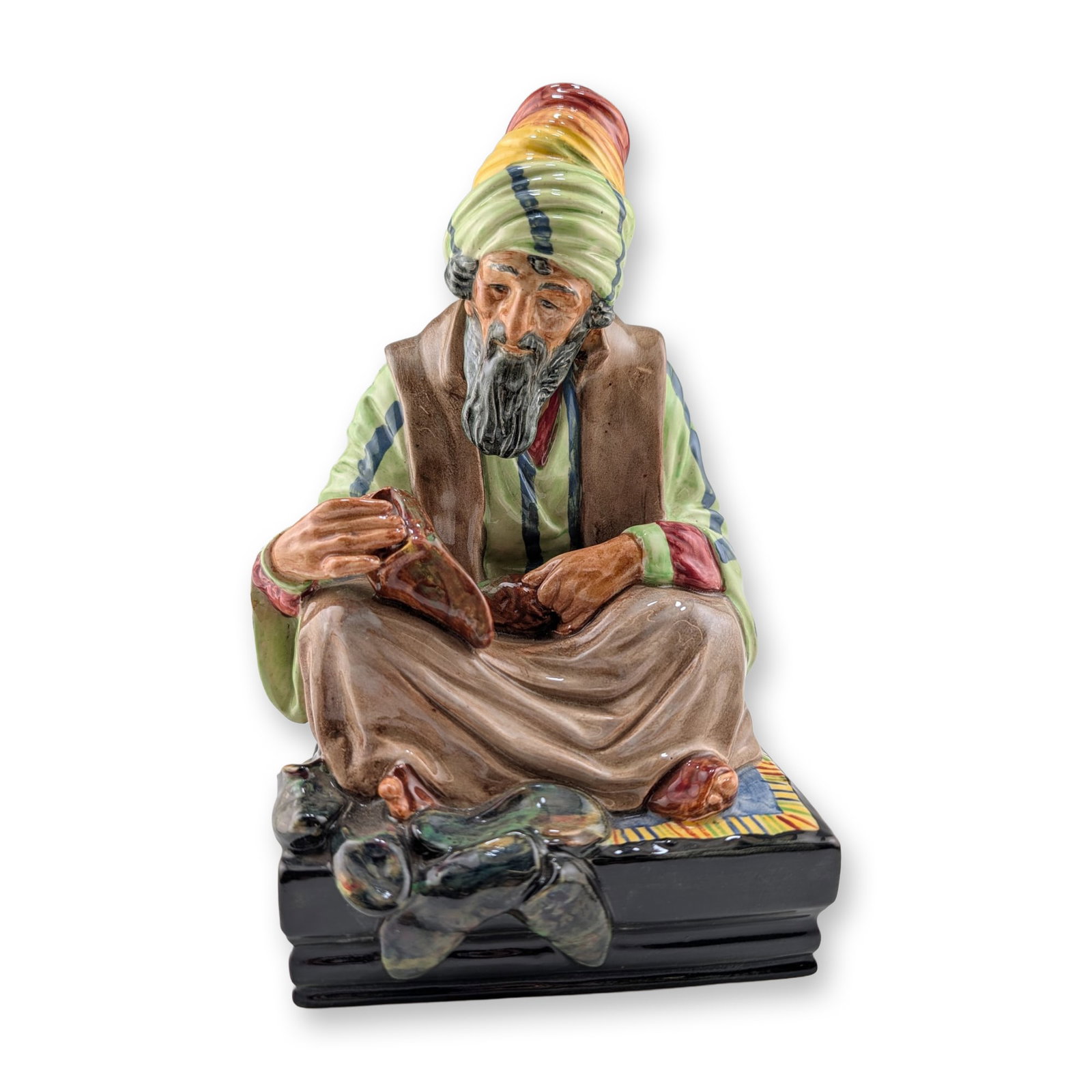 Royal Doulton Ceramic Figurine, Cobbler HN1706: Glazed ceramic figurine depicting a bearded man in traditional Middle Eastern attire seated cross-legged on a patterned rug. The figure is dressed in a striped green and white robe, brown vest, and a 