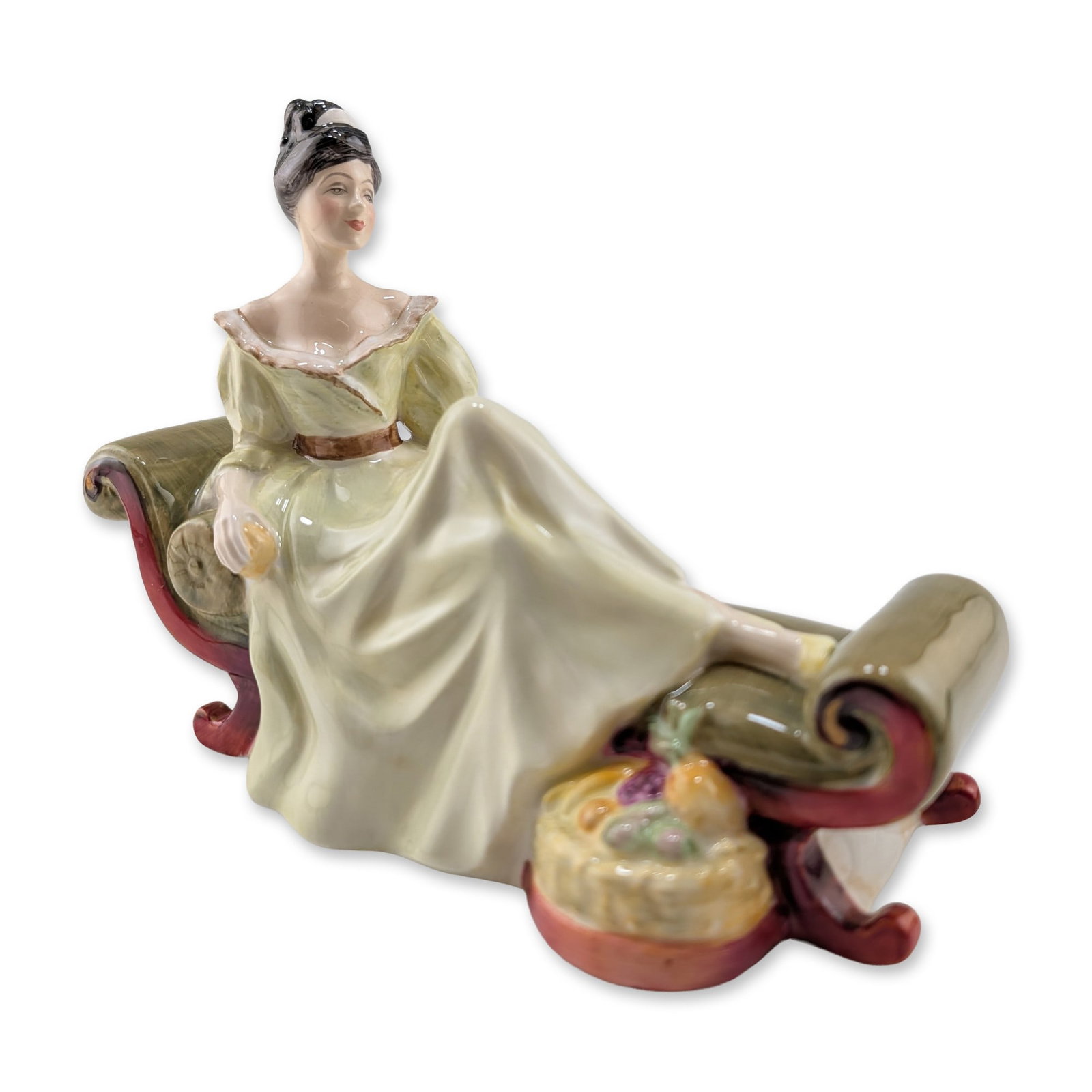 Royal Doulton Bone China Figurine, At Ease HN2473: The piece depicts a woman with dark hair in a formal updo, dressed in a pale yellowish-green gown with a ruffled white neckline and a brown waist sash. She is portrayed reclining on a classical chaise