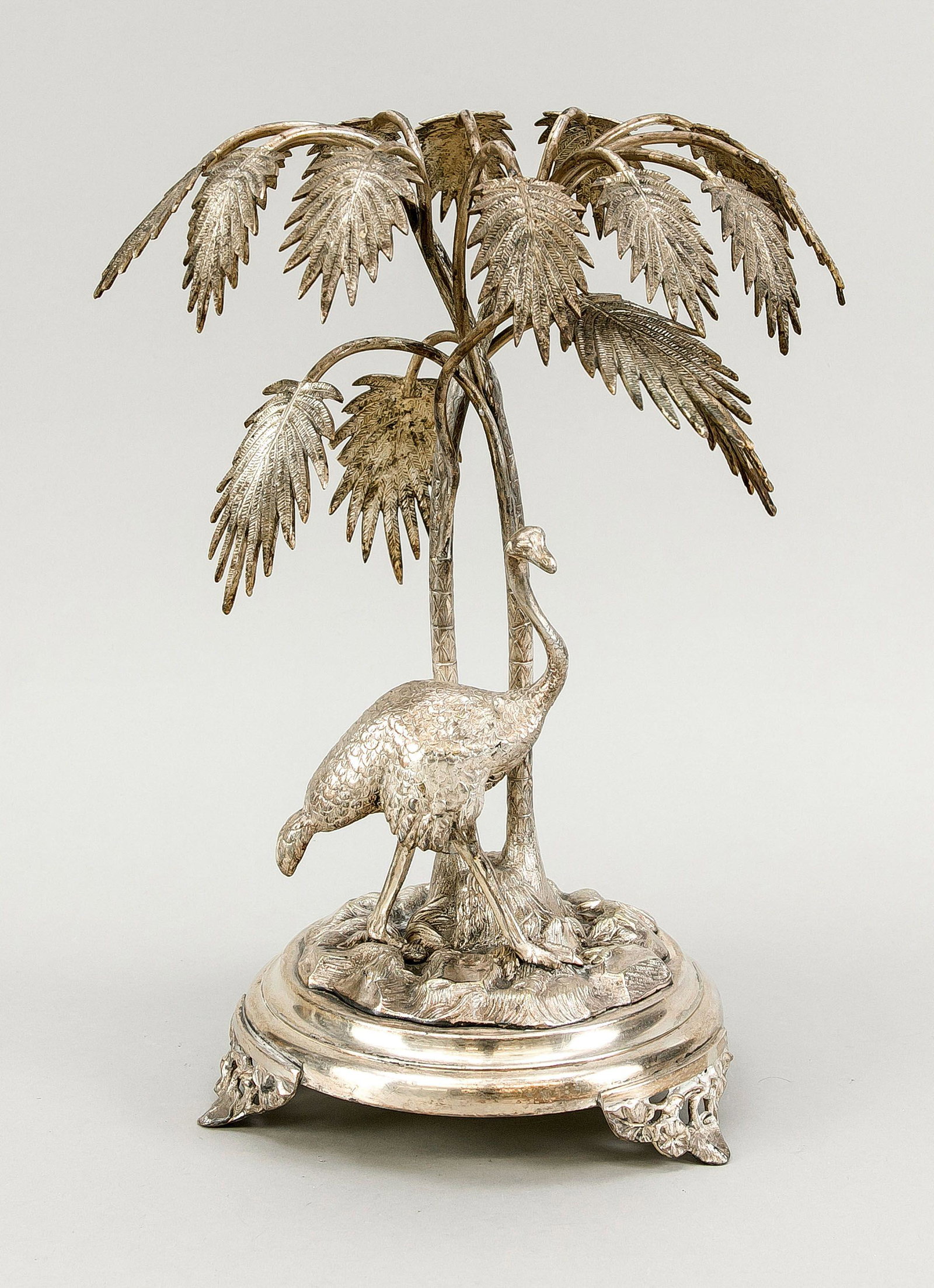 Table decoration, around 1900, meta: Table decoration, around 1900, metal, silver plated. Round base on 3 feet, on it a terrain plinth with bird ostrich and 2 palm trees. Slightly rubbed & bumped, h. 36 cm