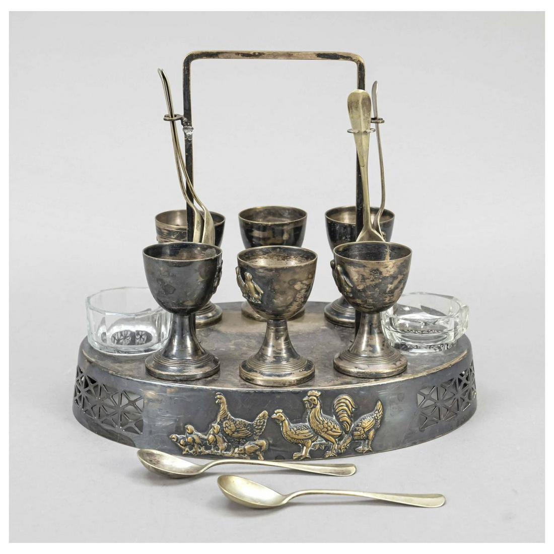 Egg cup set, early 20th c., plated, o: Egg cup set, early 20th c., plated, oval stand with central handle, wall with figural relief decoration, complete with 6 egg cups, 2 glass almonds (bumped) and 6 spoons l. 22 cm
