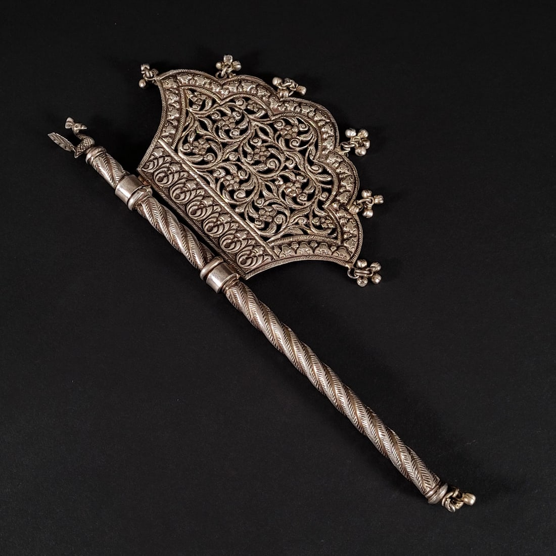 Indian Silver Mandir Temple Fan: Indian silver mandir temple fan. The ax-shaped fan features a twisted handle topped by a quintessential Indian peacock finial. The fan has vegetal openwork and is accented with hanging appliques along