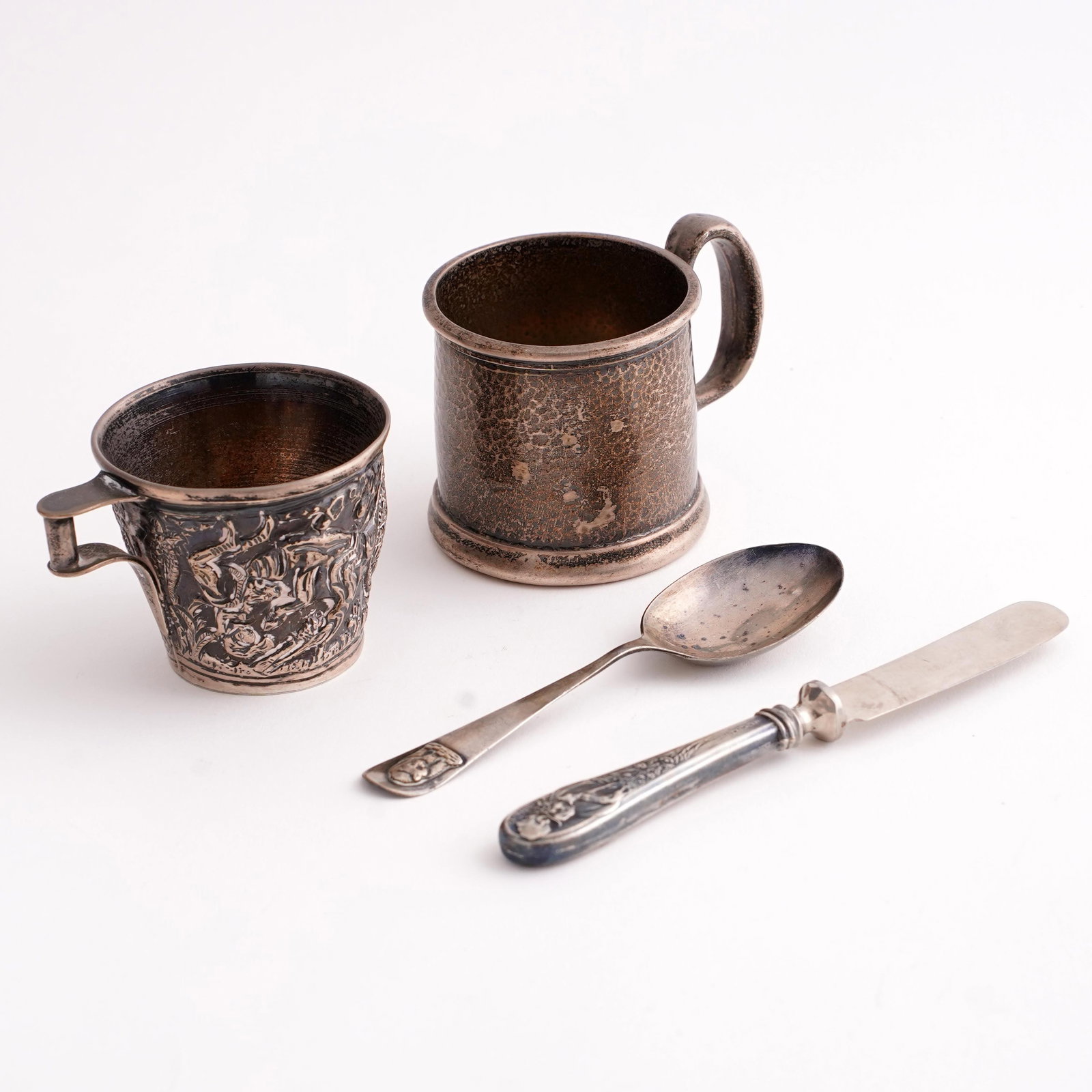 Group of Sterling Silver Table Articles: A group of sterling silver table articles comprising two small cups or cup holders, a spoon, and a handled butter spreader/knife. The smaller cup with embossed figural and foliate decoration, marked �