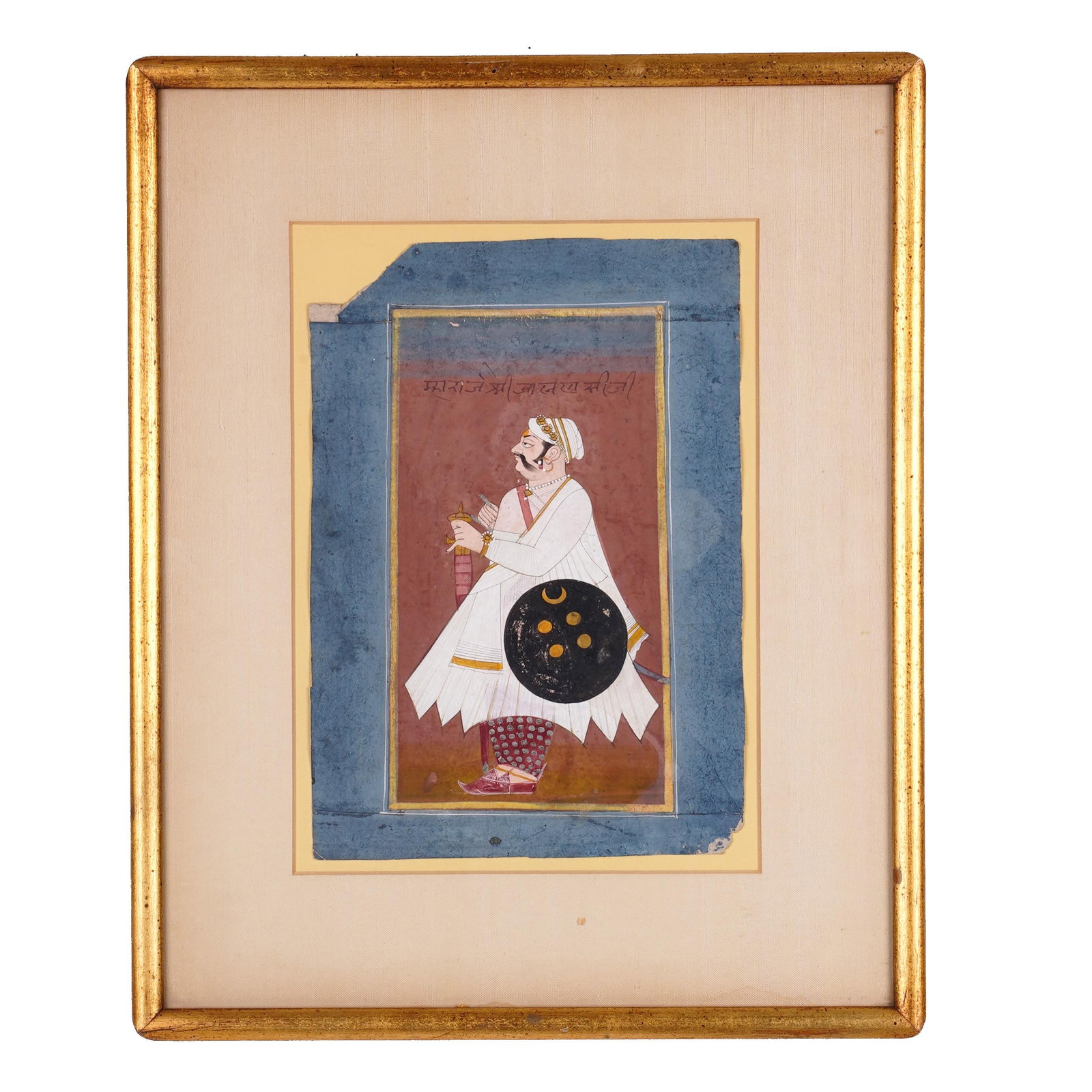 Framed Indian Miniature Painting of a Nobleman: An Indian miniature painting depicting a standing nobleman or warrior in profile, wearing white robes and jeweled turban, holding a small vessel and carrying a black shield with crescent and roundel d