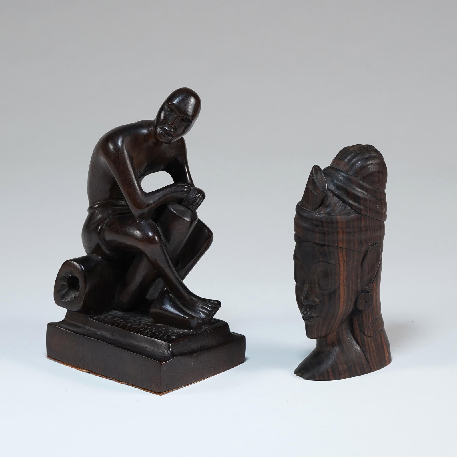 Two Carved Wood Figural Sculptures: A lot of two carved wood figural sculptures, comprising a seated male figure posed on a square plinth beside a drum-like object, and a carved female bust or head with high coiffure and stylized facial