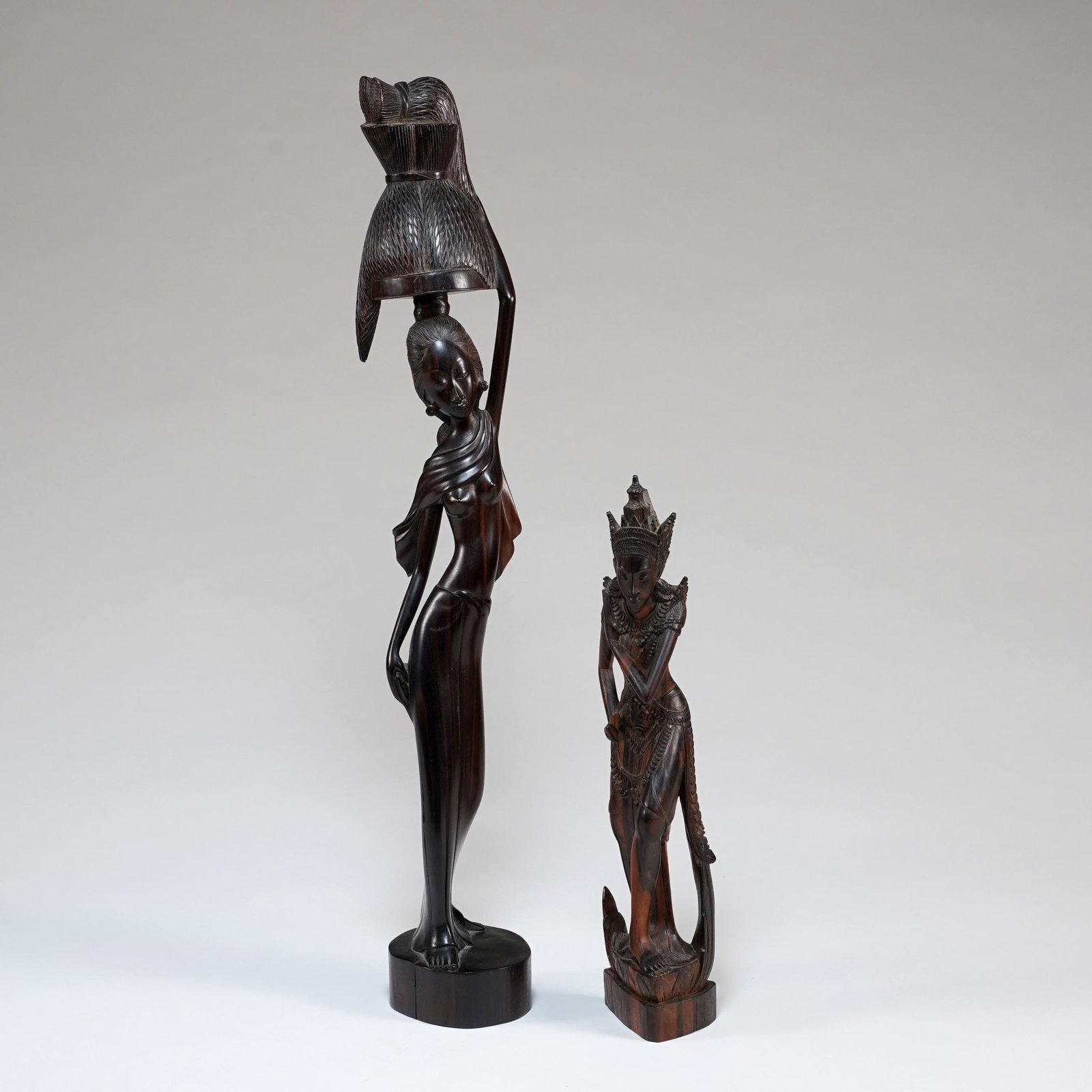 Two Southeast Asian Carved Wood Figural Sculptures: A lot of two carved wood figures, comprising a tall female figure balancing a covered basket or vessel on her head, raised on a round plinth, and a smaller elaborately carved standing dancer or deity 