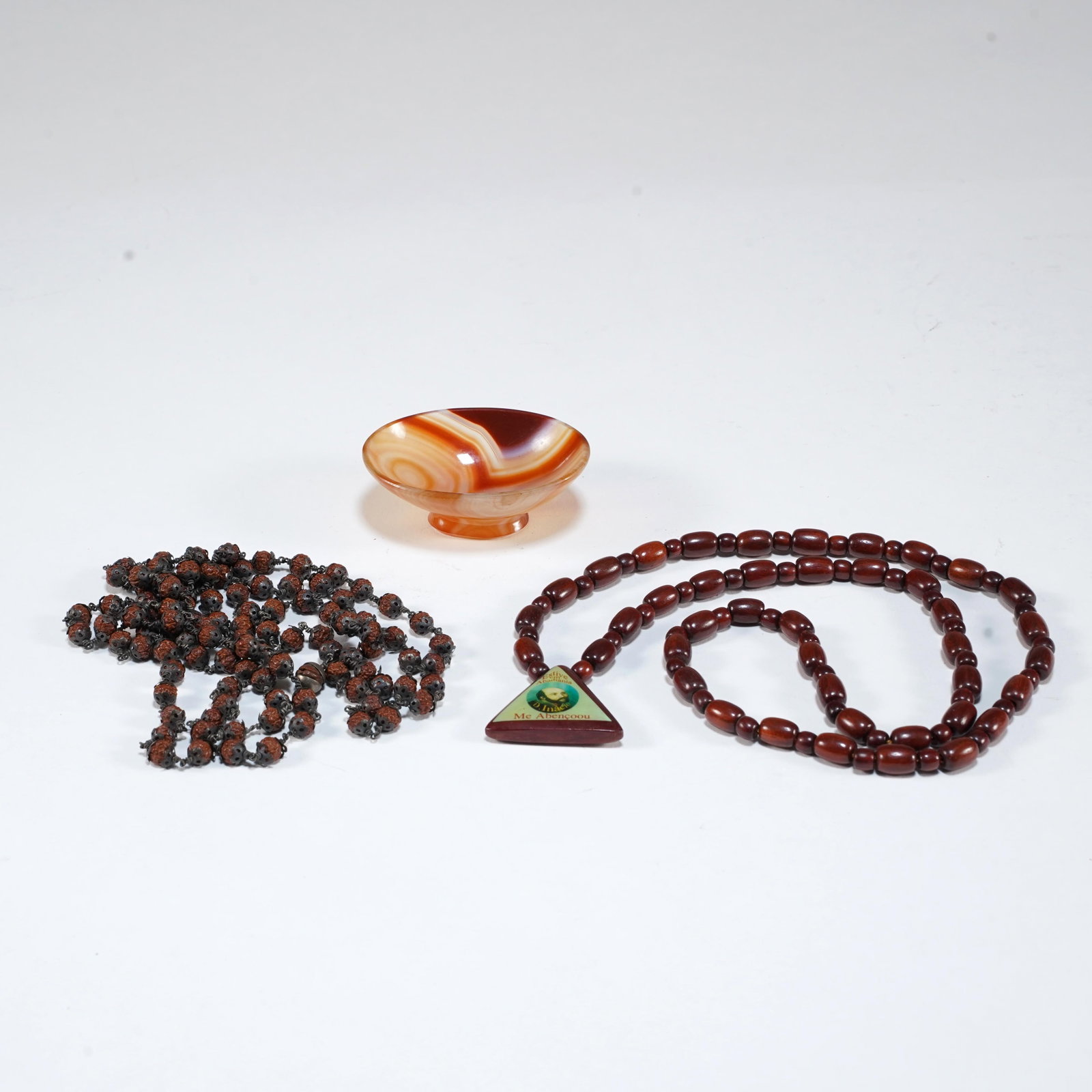 Group of Beads and Agate Bowl: A small decorative group comprising a polished banded agate footed bowl, a strand of dark rudraksha-style prayer beads with metal spacer elements, and a reddish-brown wooden or seed-bead necklace