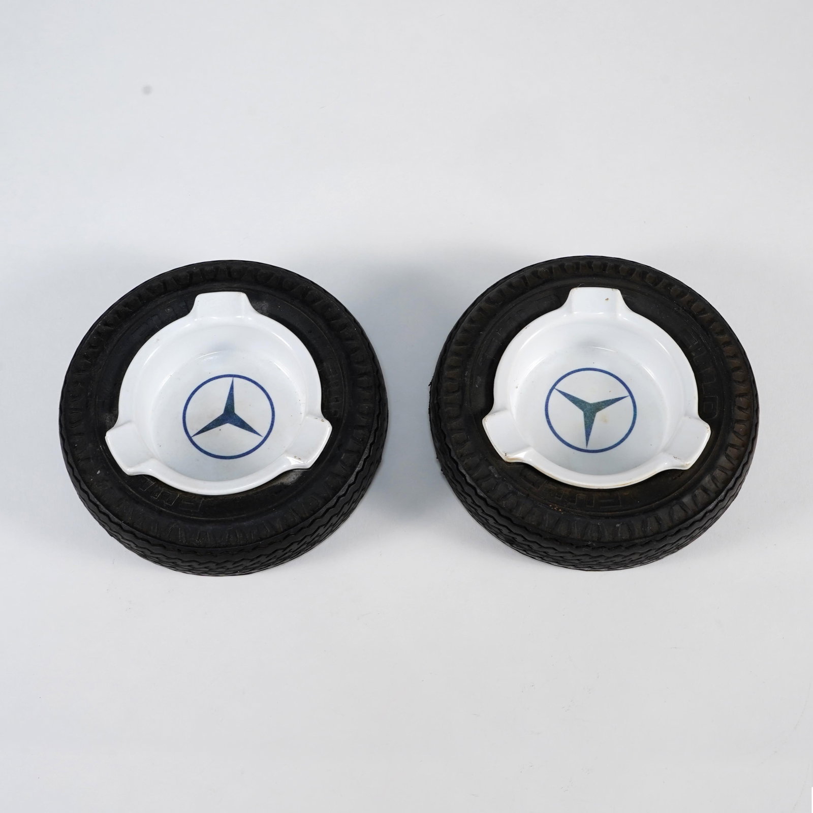 Pair of Mercedes-Benz Tire-Form Ashtrays: Pair of Mercedes-Benz tire-form ashtrays, each with white ceramic ashtray insert bearing the blue three-pointed star emblem, set within black rubber tire-form surround with molded tread decoration.