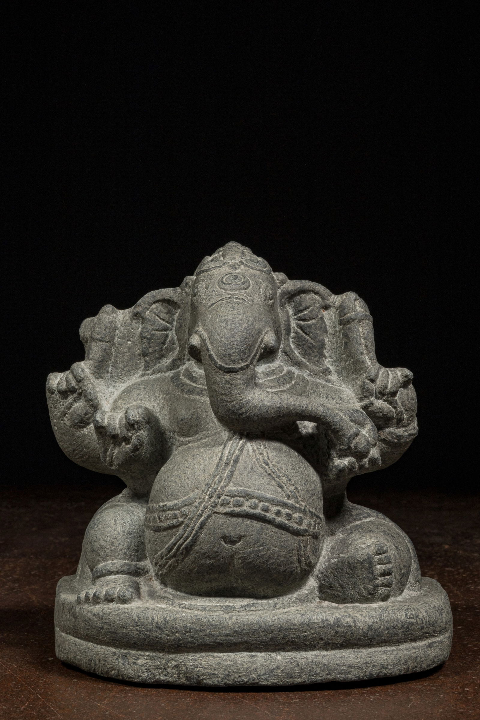 SEATED STONE GANESHA INDIA 12TH CENTURY OR LATER: Dimensions: Height 17,5 cm Wide 16 cm Depth 10 cm Weight: 3362 grams A finely carved stone sculpture of the Hindu deity Ganesha, depicted in a seated posture. The figure is rendered in dark grey stone