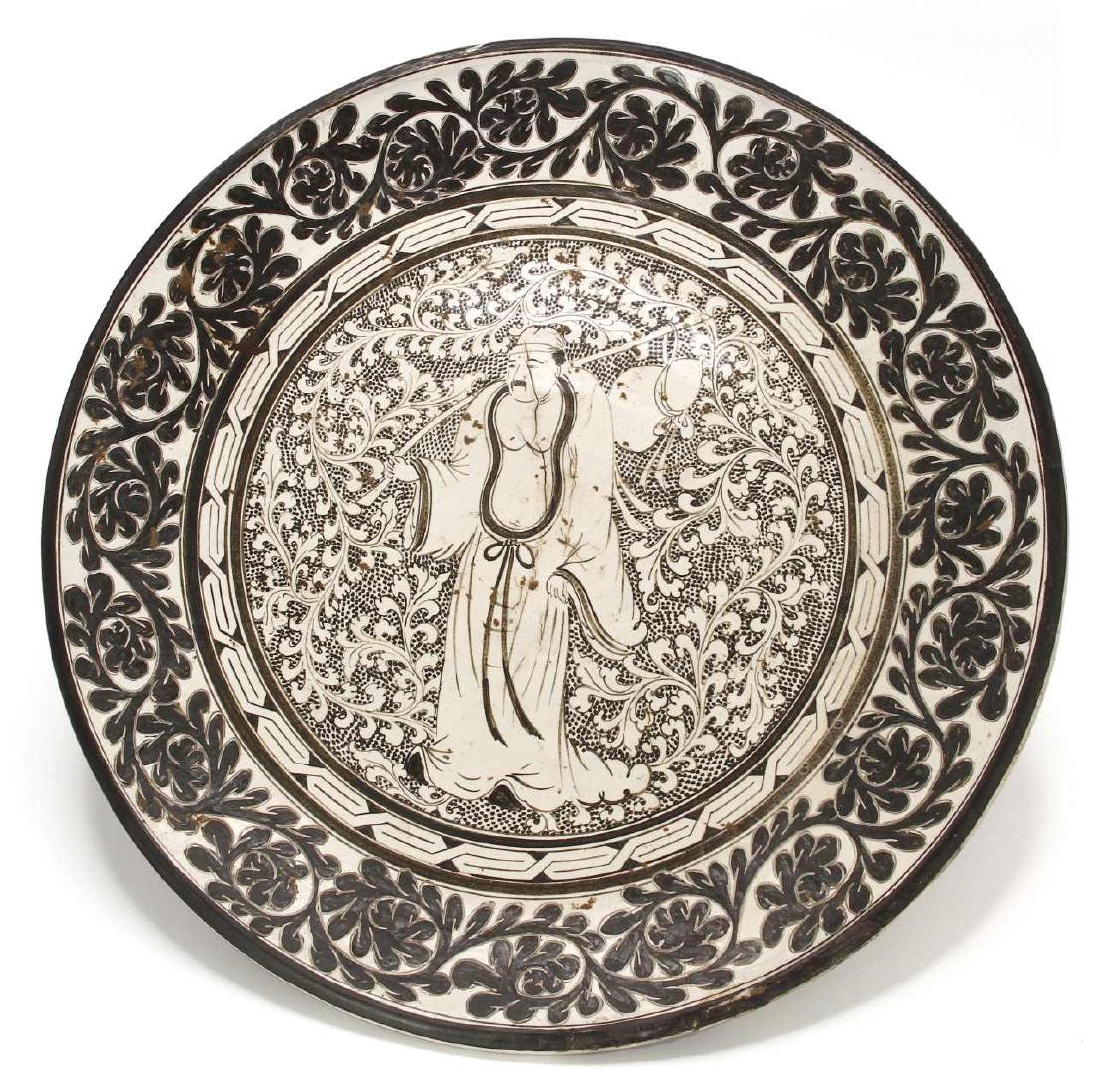 Large T'su-Chou Sagraffiato Dish: The thickly potted dish with a center medallion decorated in a dark brown slip, painted with a Daoist figure on a scrolling floral ground, and encircled by a band of meandering floral sprigs carved th