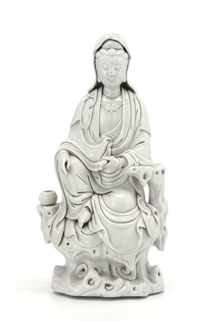 Dehua Figure of Guanyin: Portrayed seated on rockwork supporting a bowl, holding a scroll, wearing an elegantly draped robe extending to a cowl covering the chignon, adorned only with bangles encircling the wrists of the sepa