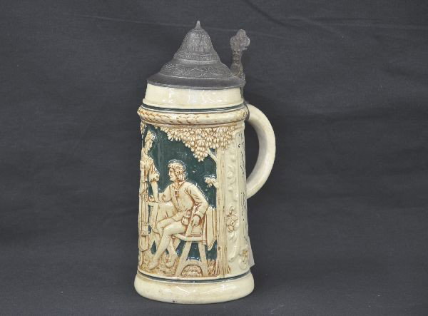 Mettlach Beer Stein: 9" tall ceramic with tin top, reads "Under Beutel Schlaff U Leer" on one side "Keimen Tropfen im Bedner Mehr" on other side. Clean, finish has light crazing.