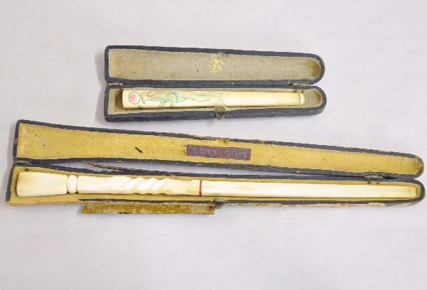 Carved Ivory Cigarette Holders: Includes a 9 1/8" Art deco holder with amber ring, hard case worn; 4 1/2" hand tinted lightly carved ivory with eagle headed figure holding leaf motif with hard case. Both show signs of use & age. Exa