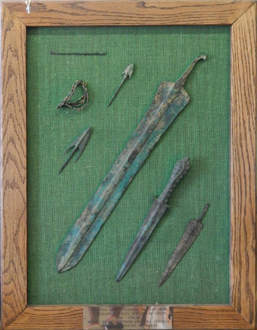 7 ANCIENT WEAPONS TOOLS MUMMY BEADS WALL DISPLAY: A collection of 7 ancient weapons and tools set on a wall display frame. Lower center of frame holds plaque that states "Weapons are from battlefield pick-ups from Luristanian (Persia) invasion of Egy