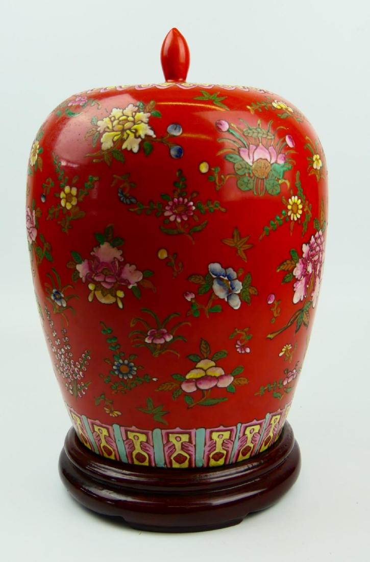 CHINESE FAMILLE ROSE FLORAL GINGER JAR VASE: Chinese hand painted Famille Rose enameled covered vase having a floral bloom design over rust red ground. Bottom holds red six character zhuanshu Qianlong reign mark. Includes fitted wooden base. Mea