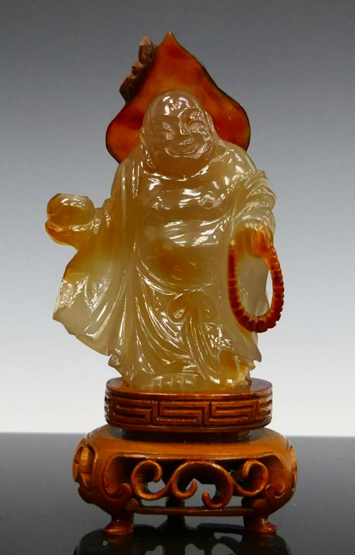 CHINESE CARVED CARNELIAN AGATE HOTIE BUDDHA FIGURE: A Chinese carved carnelian agate stone sculpture depicting standing Hotei Buddha with beaded necklace and jar. Includes fitted wooden display stand. Measures approx. 5" height (12.7cm) overall includi