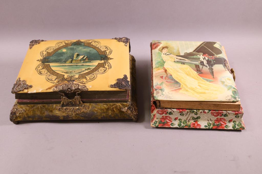 TWO VICTORIAN PHOTO ALBUM MUSIC BOXES: CELLULOID SCENIC LIDS, PHOTO ALBUMS WITH MUSIC BOX BASE. LATCH IS BROKEN ON ONE. DAMAGE TO ONE LID. 11 1/2" - 12 1/2"