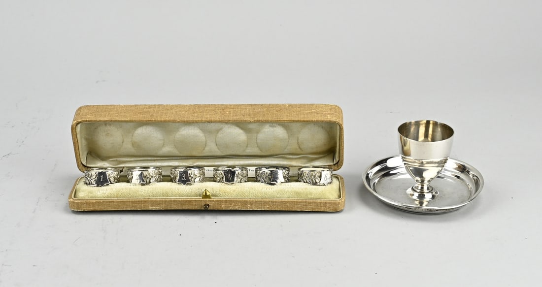 Silver egg cup and finger cloth rings: Silver 6 finger cloth rings, 800/000, in cassette and an egg cup, 833/000. Six openwork finger cloth rings with oak leaves and a shield with a number. ø 26x11mm. And an egg cup attached to a round di