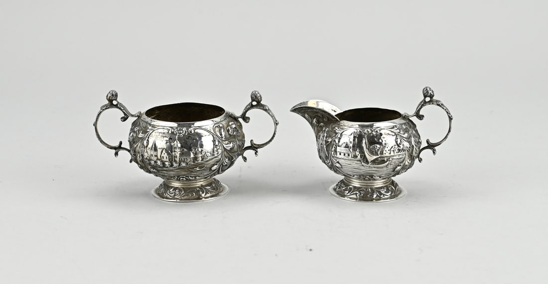 Silver milk and sugar bowl: Small silver milk and sugar bowl, 833/000, round model with floral decoration and representations of a water with a cityscape. The handles are provided with putti heads. MT.: G.Schoorl, Zaandijk,