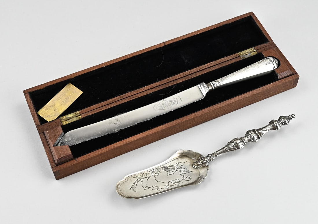 Cassette with bread knife & pastry scoop: Cassette with a bread knife with a silver handle with hammer blow decoration and floral elements. Frans Brugsma Groningen, 31 cm. and a cake shovel with contoured handle. Silver-containing alloy. In g