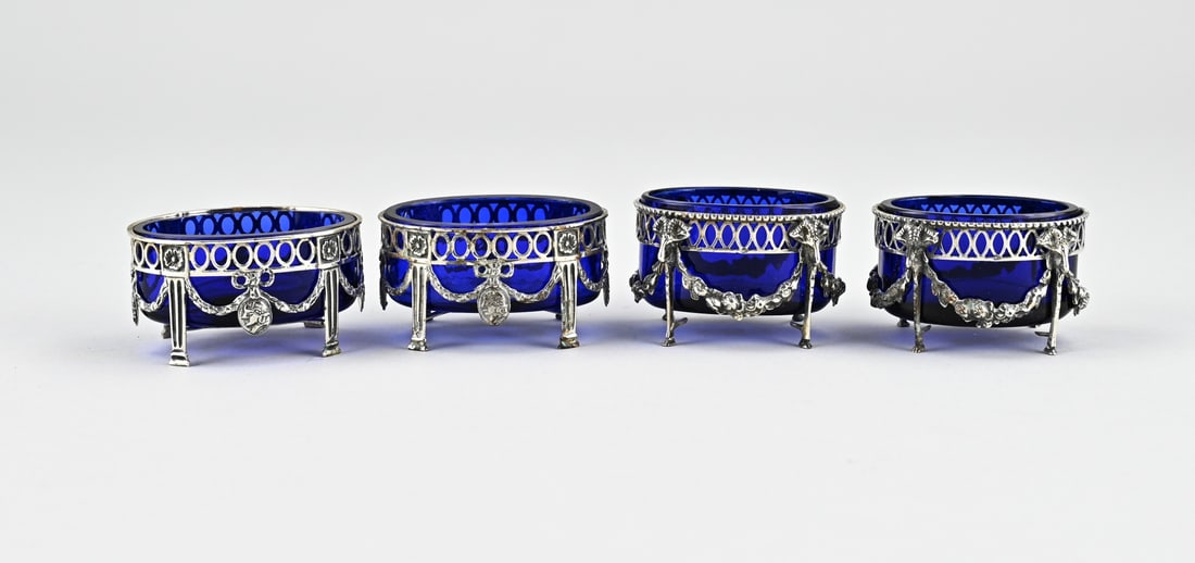 Silver salt cellars in frame: Four silver spice bowls, 833/000, with blue glass. Two oval bowls decorated with garlands and medallions and two oval bowls with garlands and ram's heads. Both with an oval sawn decoration. MT.: