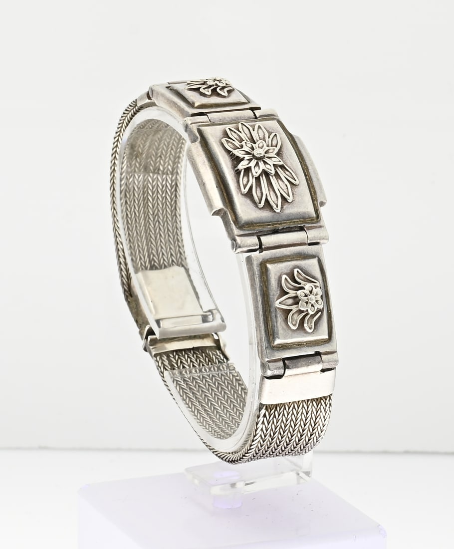 Silver bracelet: Silver bracelet, 925/000, with a partly braided band and in the middle 3 rectangular elements provided with a flower element. Provided with a box clasp with safety. Length 18.5 cm. Width 20 mm. In