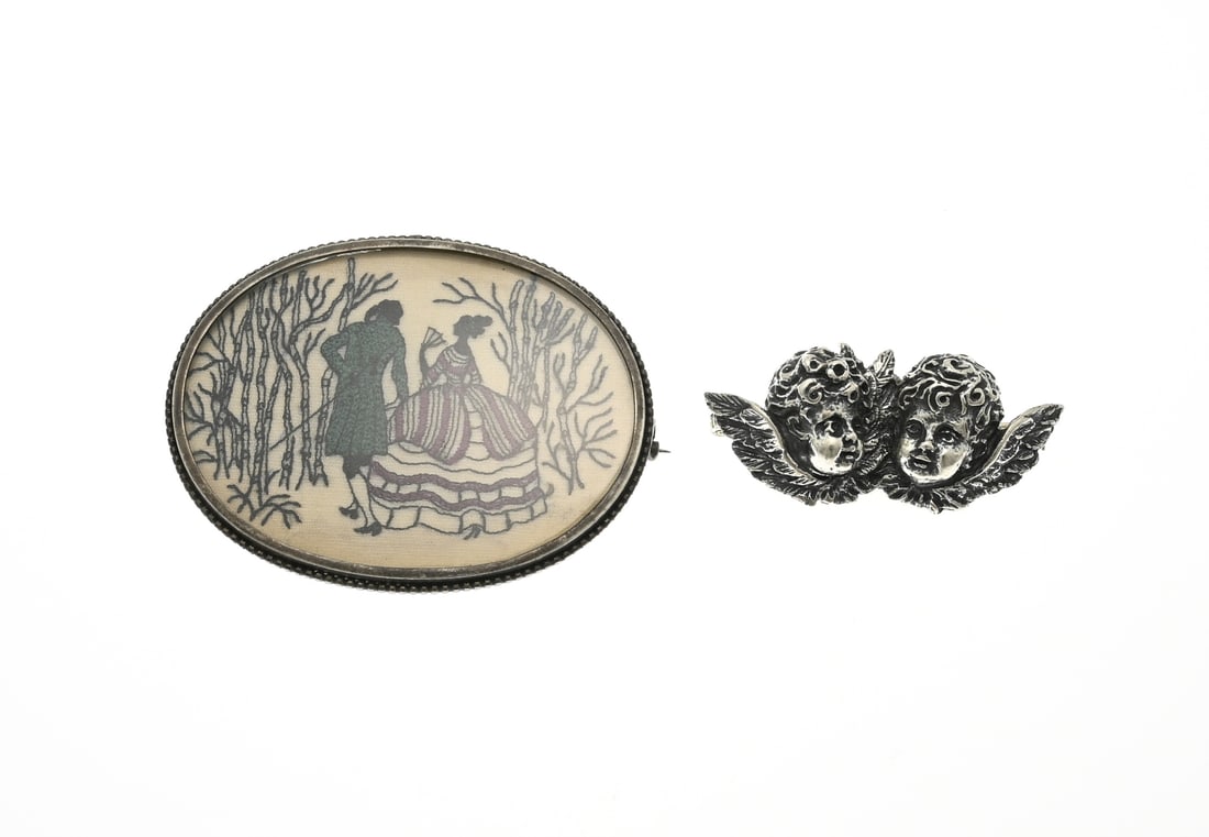 2 Brooches: Lot with 2 silver brooches, one in the shape of 2 putti heads, 5 cm., and an oval brooch with a silver border with an embroidered representation of a nobleman and woman. 6x5cm. In good condition.