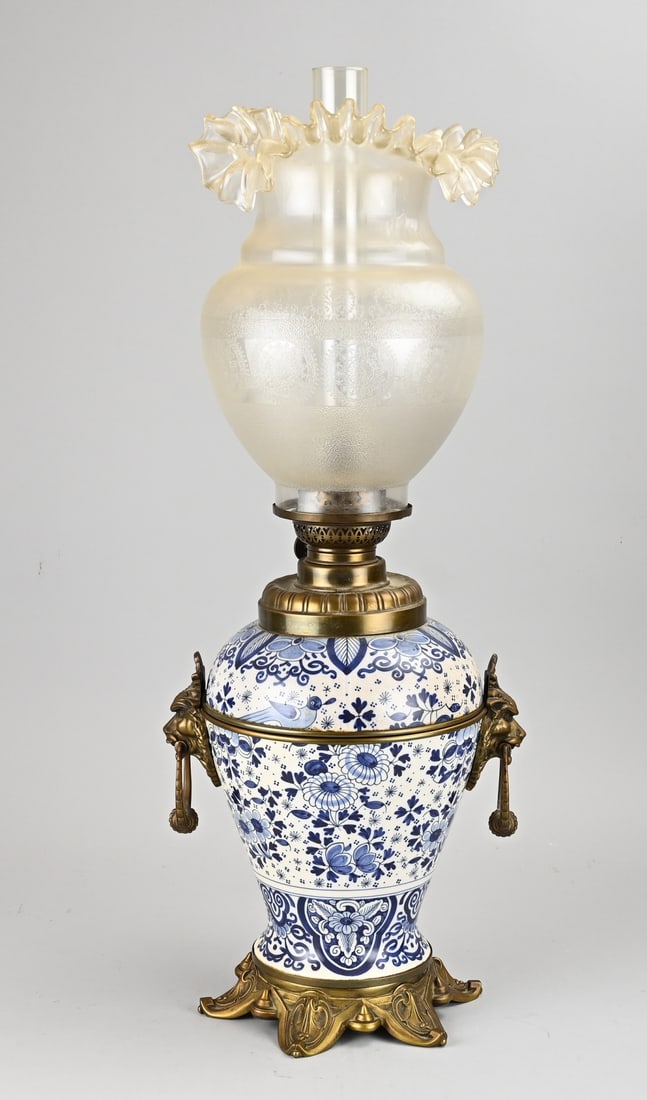 Delft kerosene lamp, 1900: Large Delft faience kerosene lamp with a glass shade and pleated collar and bronze lion heads. Circa 1900. Dimensions: H 61 cm. In good condition.